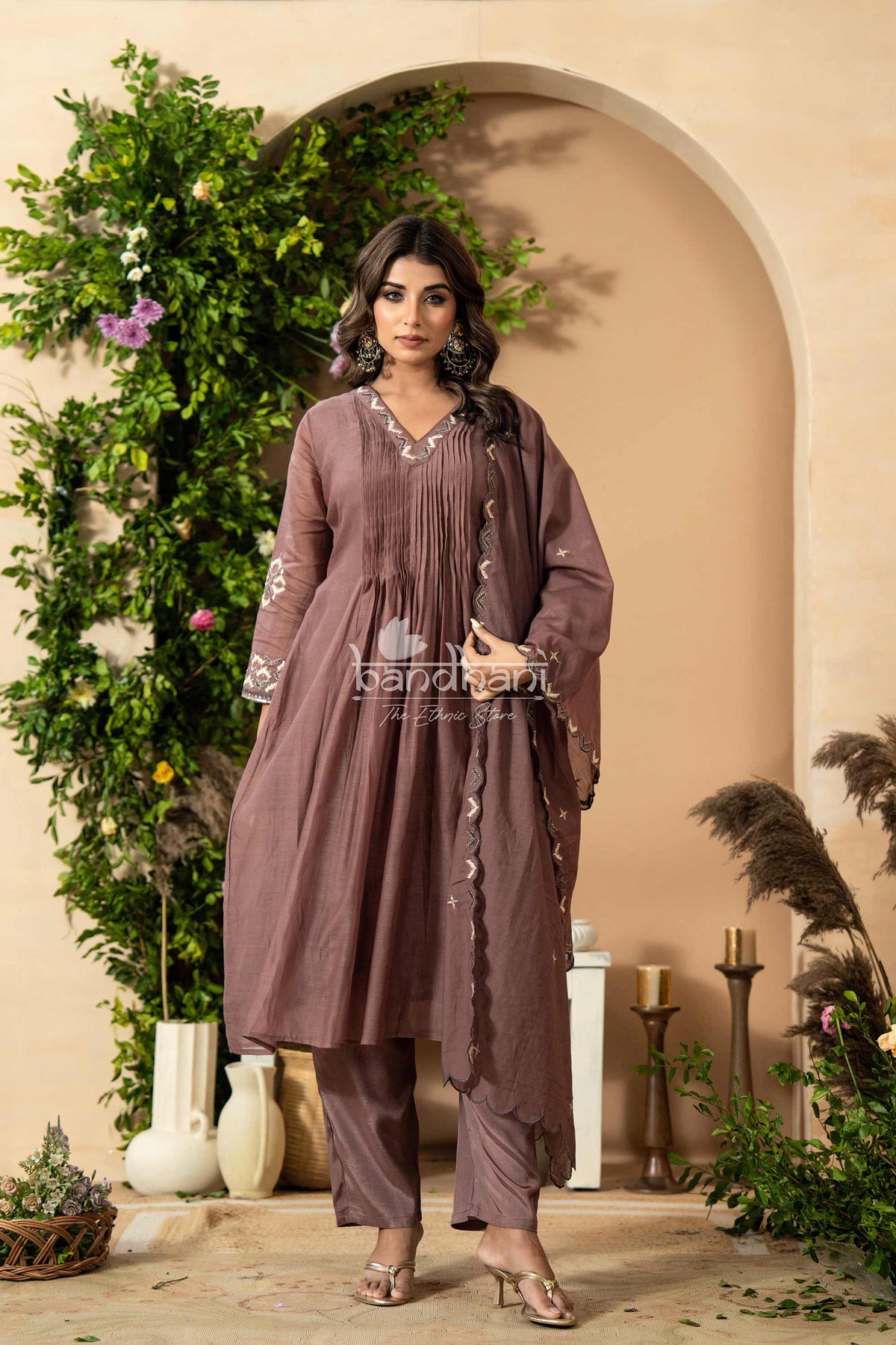 Woman in a mauve traditional outfit standing in a decorated indoor setting with plants and candles.