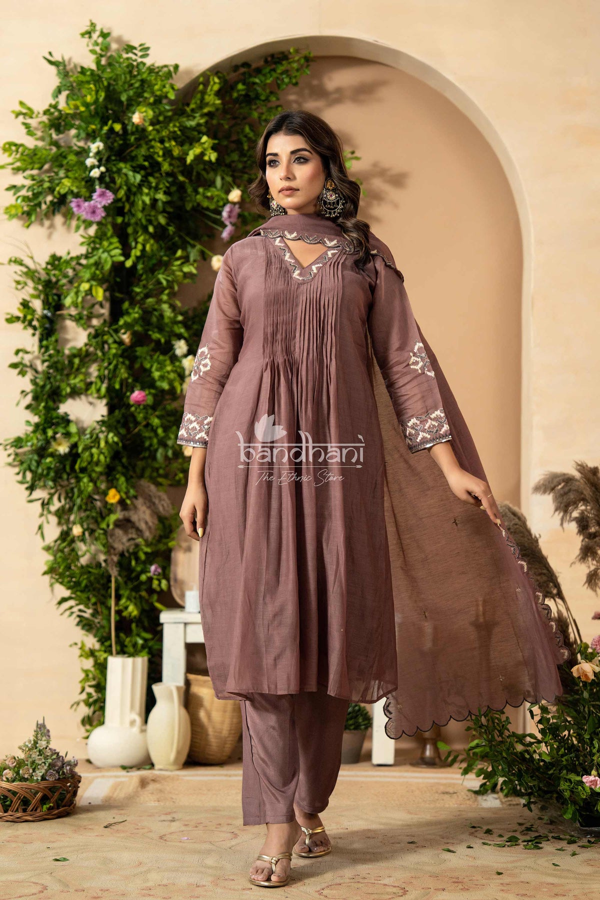 Woman wearing a brown embroidered outfit standing in front of an archway with floral decorations.