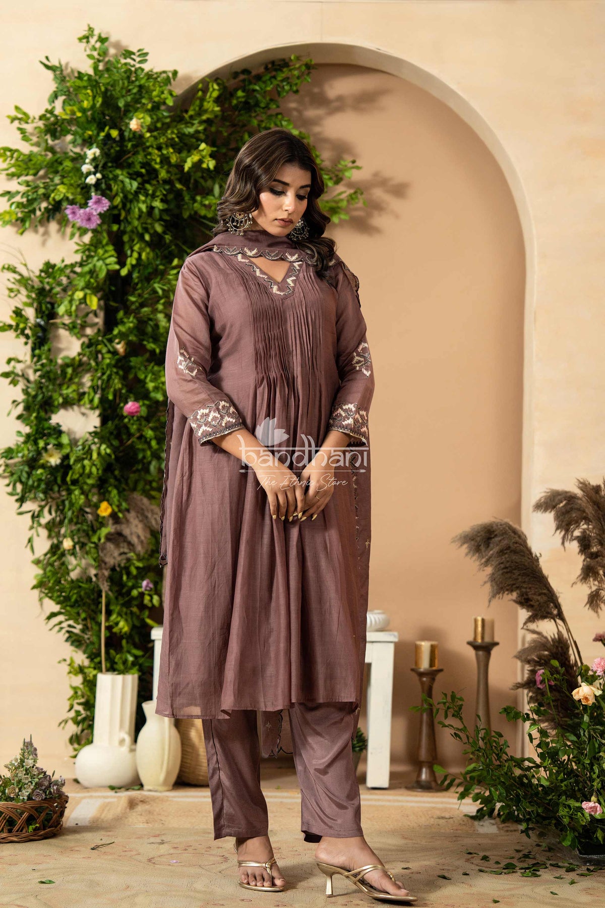 Woman in a brown traditional outfit standing in a decorated indoor setting with plants and candles.