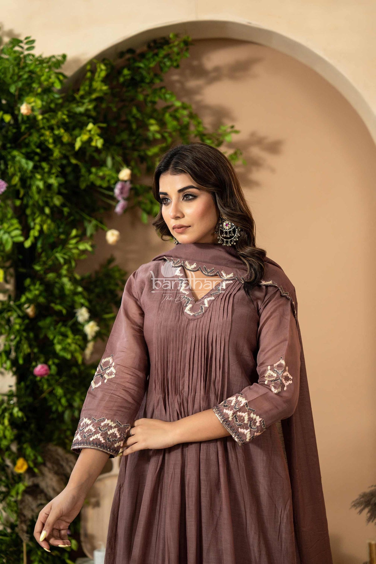 Woman wearing a brown embroidered dress with a beige wall and greenery in the background