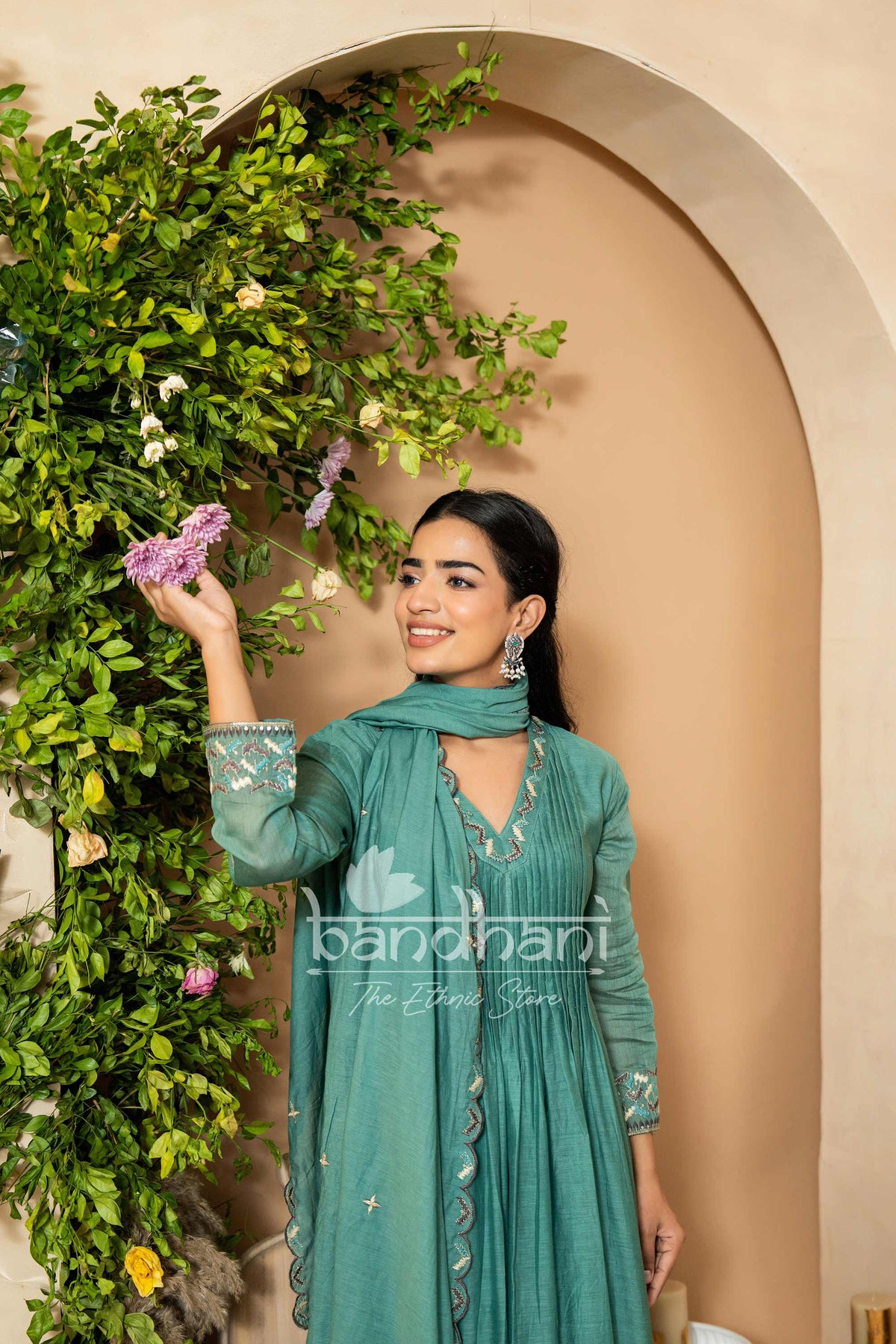 Woman in a teal outfit standing next to a decorative wall with greenery
