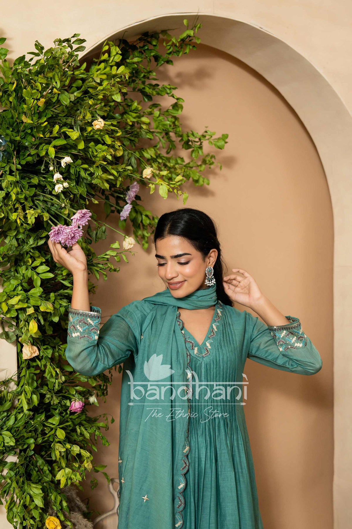 Woman in a teal traditional outfit standing next to greenery with 'bandhani' branding.