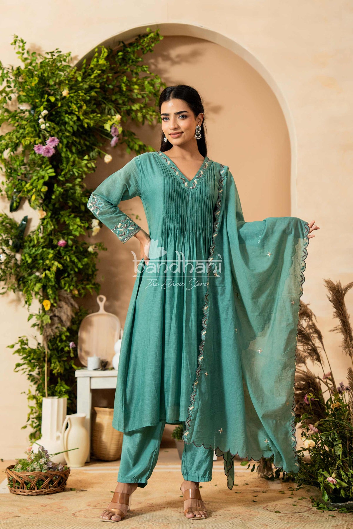 Woman in a teal traditional outfit standing in front of decorative plants and archway.