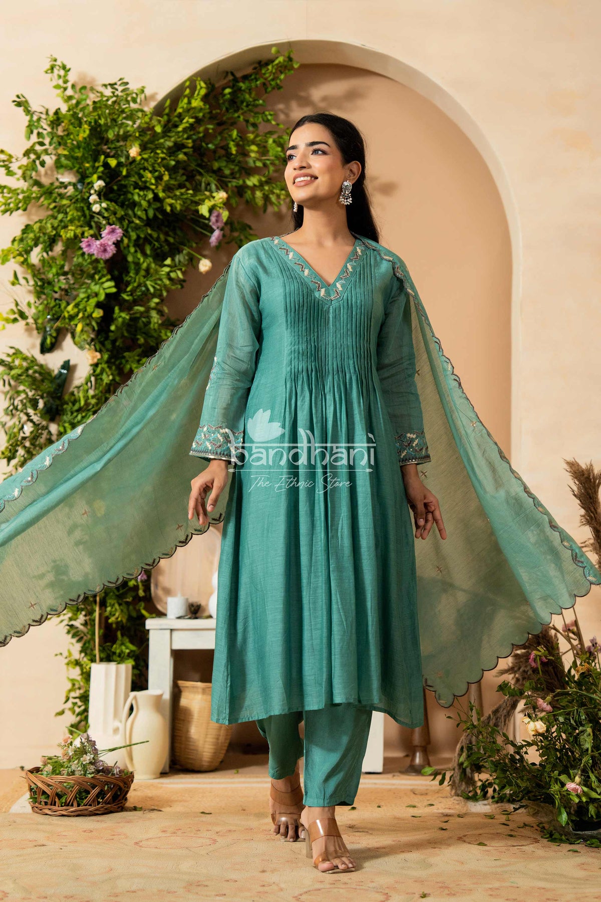 Woman in a teal traditional outfit with a flowing dupatta, standing in a decorative indoor setting.
