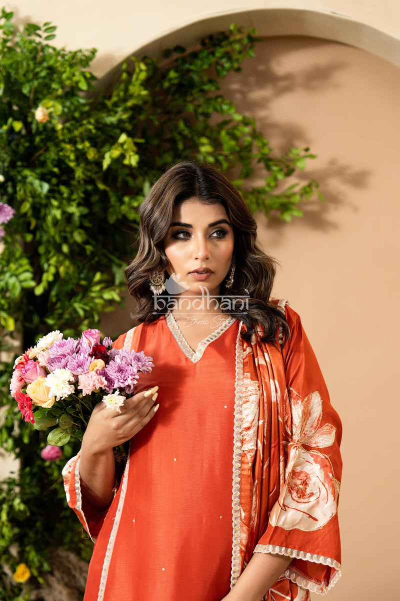 Woman in an orange outfit with floral design holding flowers, standing against a beige wall with greenery.