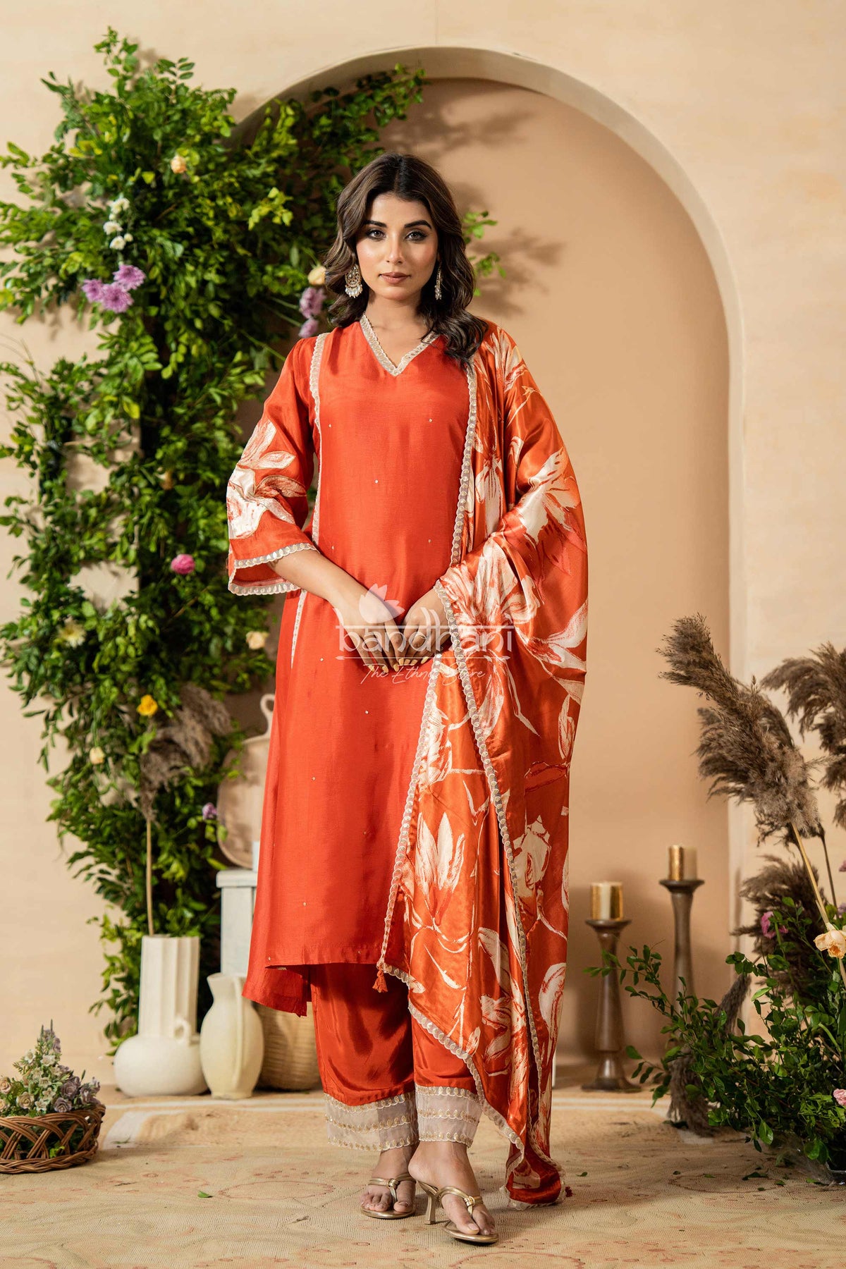 Woman in a red traditional outfit standing in a decorated indoor setting with plants and candles.