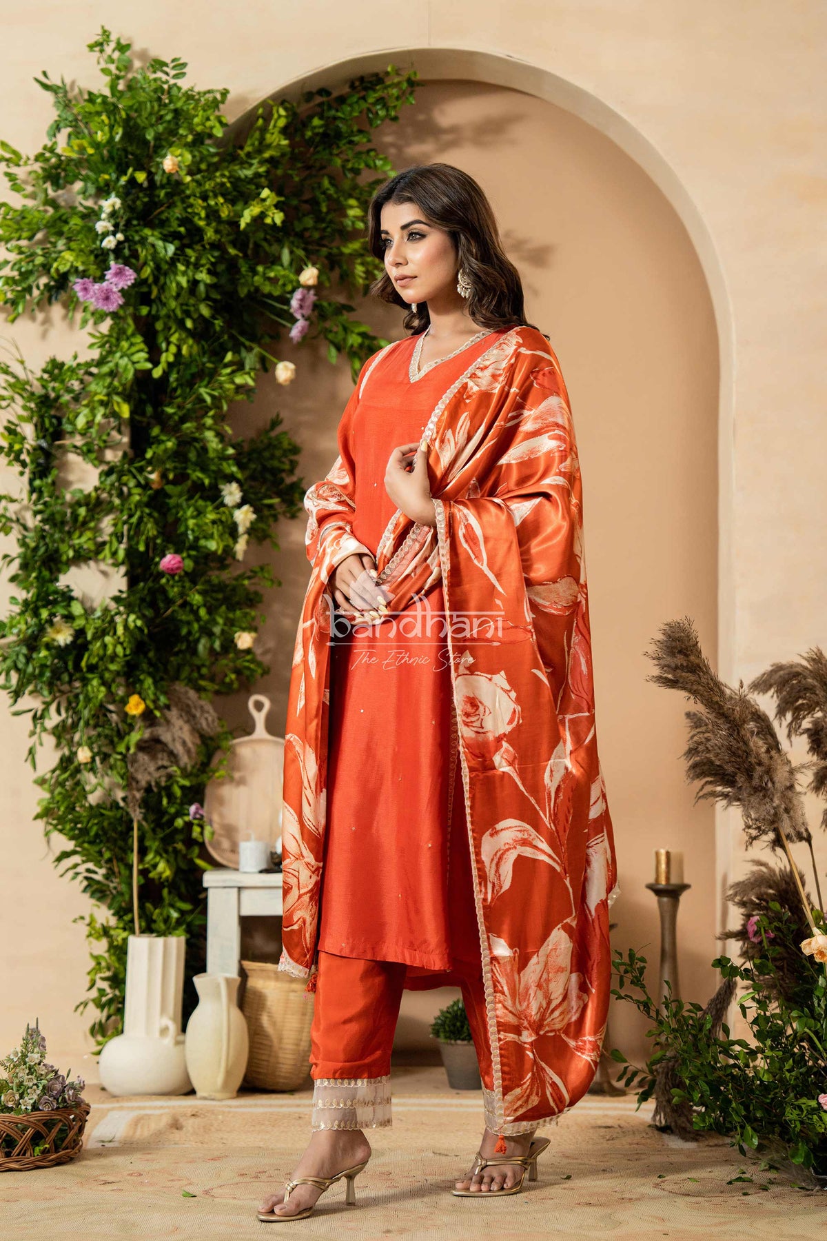 Woman in an orange traditional outfit with floral patterns standing in a decorated indoor setting.