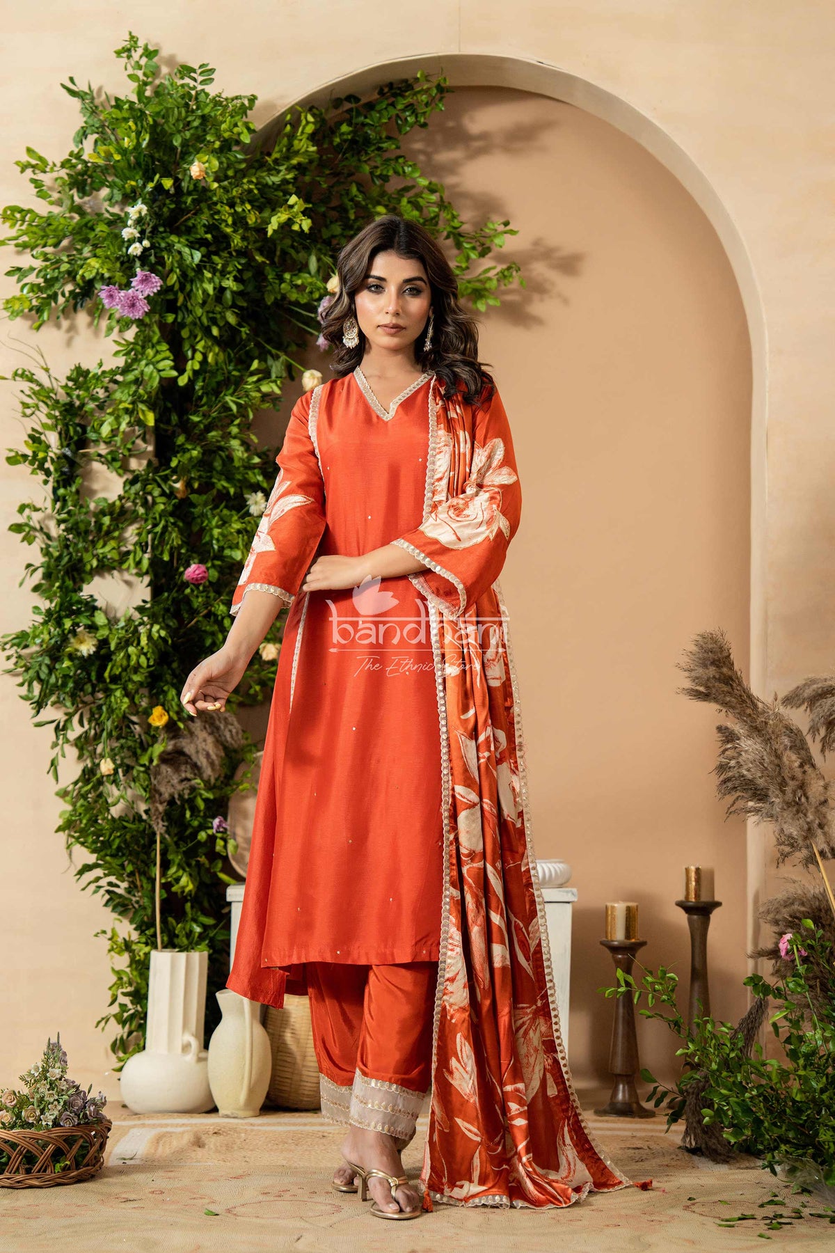 Woman in an orange traditional outfit standing in a decorated room with plants and candles.