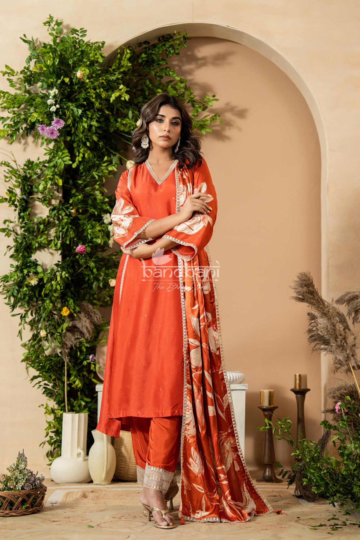 Woman in a red traditional outfit standing in a decorated indoor setting with plants and candles.