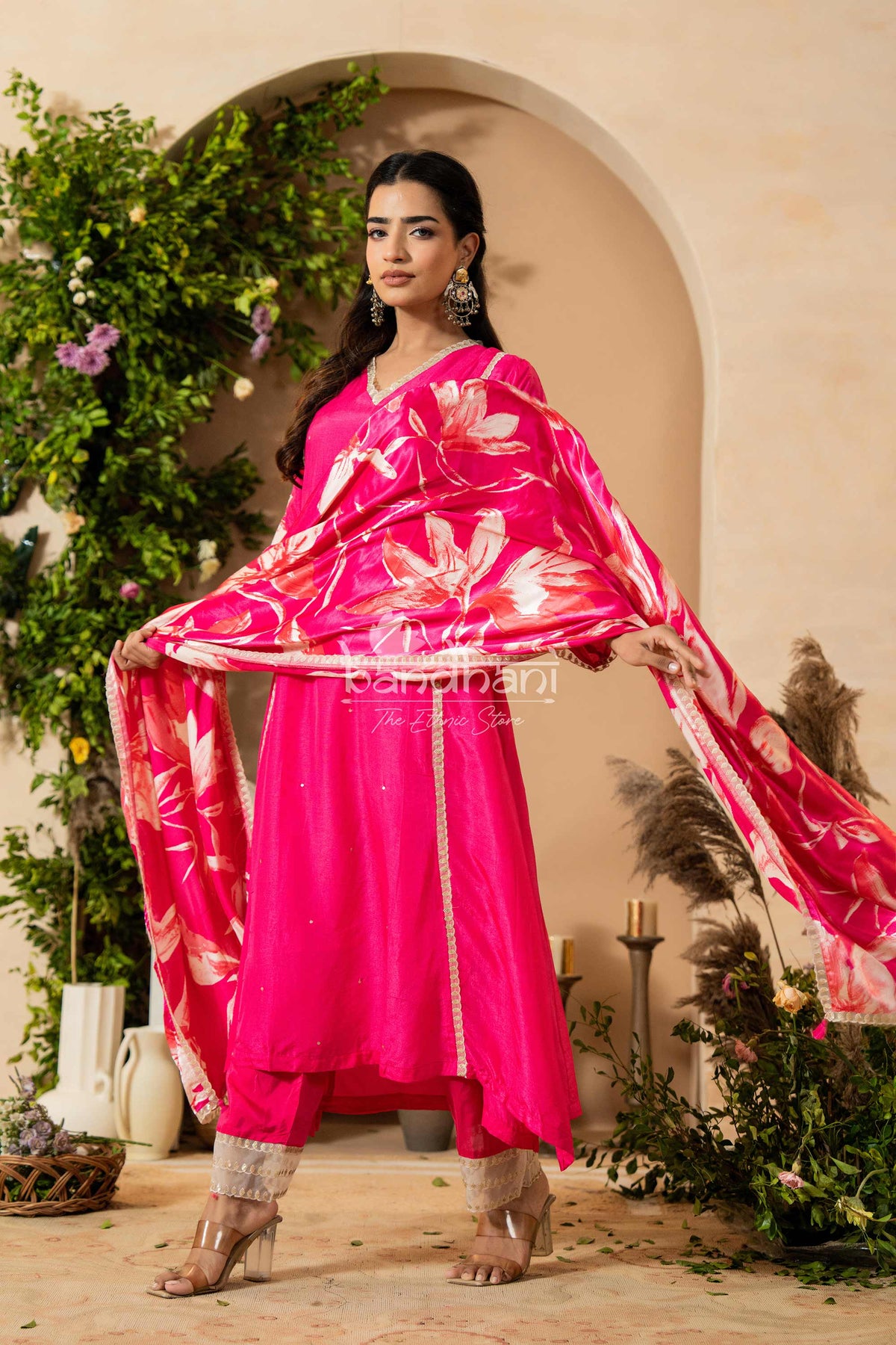 Woman in a bright pink traditional outfit with a floral dupatta, standing in a decorative setting.