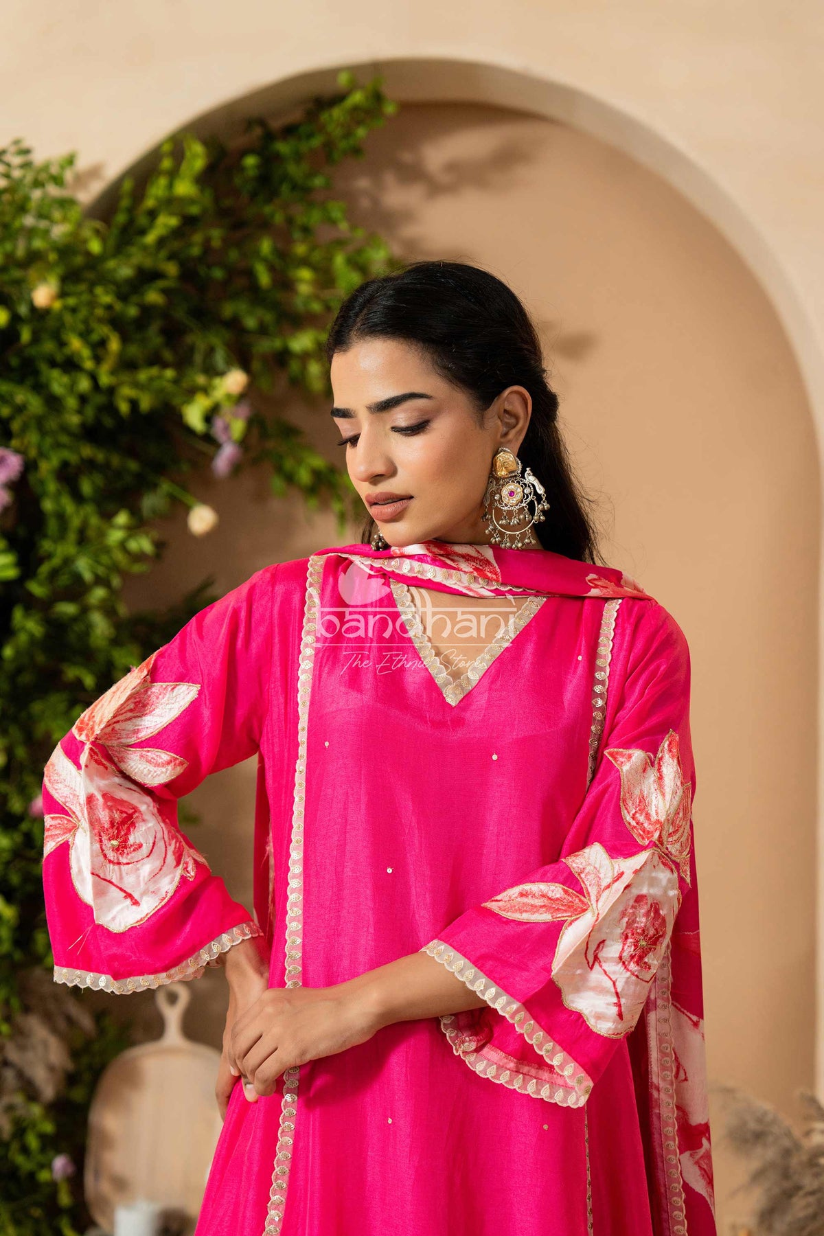 Woman wearing a bright pink traditional outfit with floral patterns, standing against a beige wall with greenery.