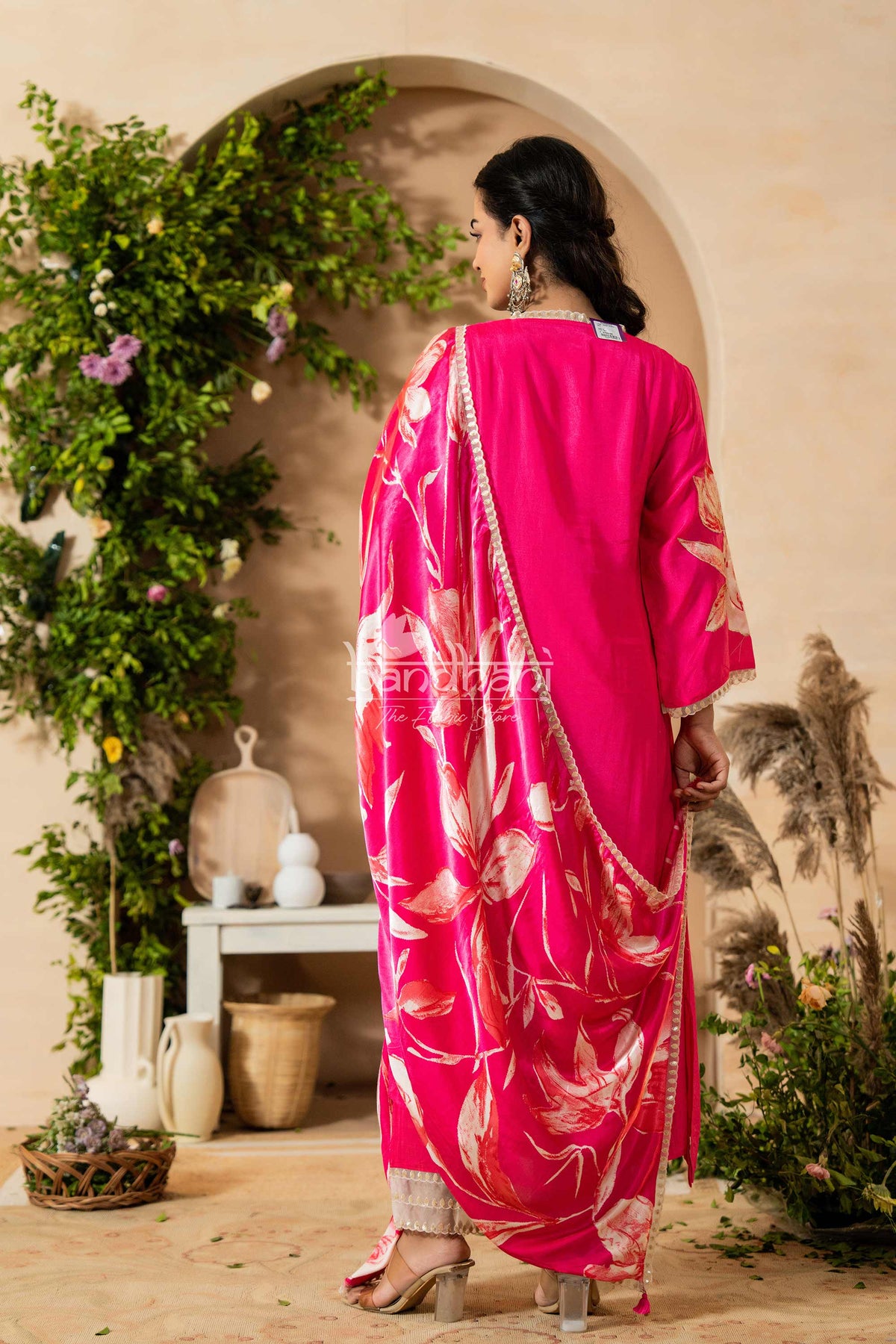 Woman in a bright pink traditional outfit with floral patterns standing in a decorative indoor setting.