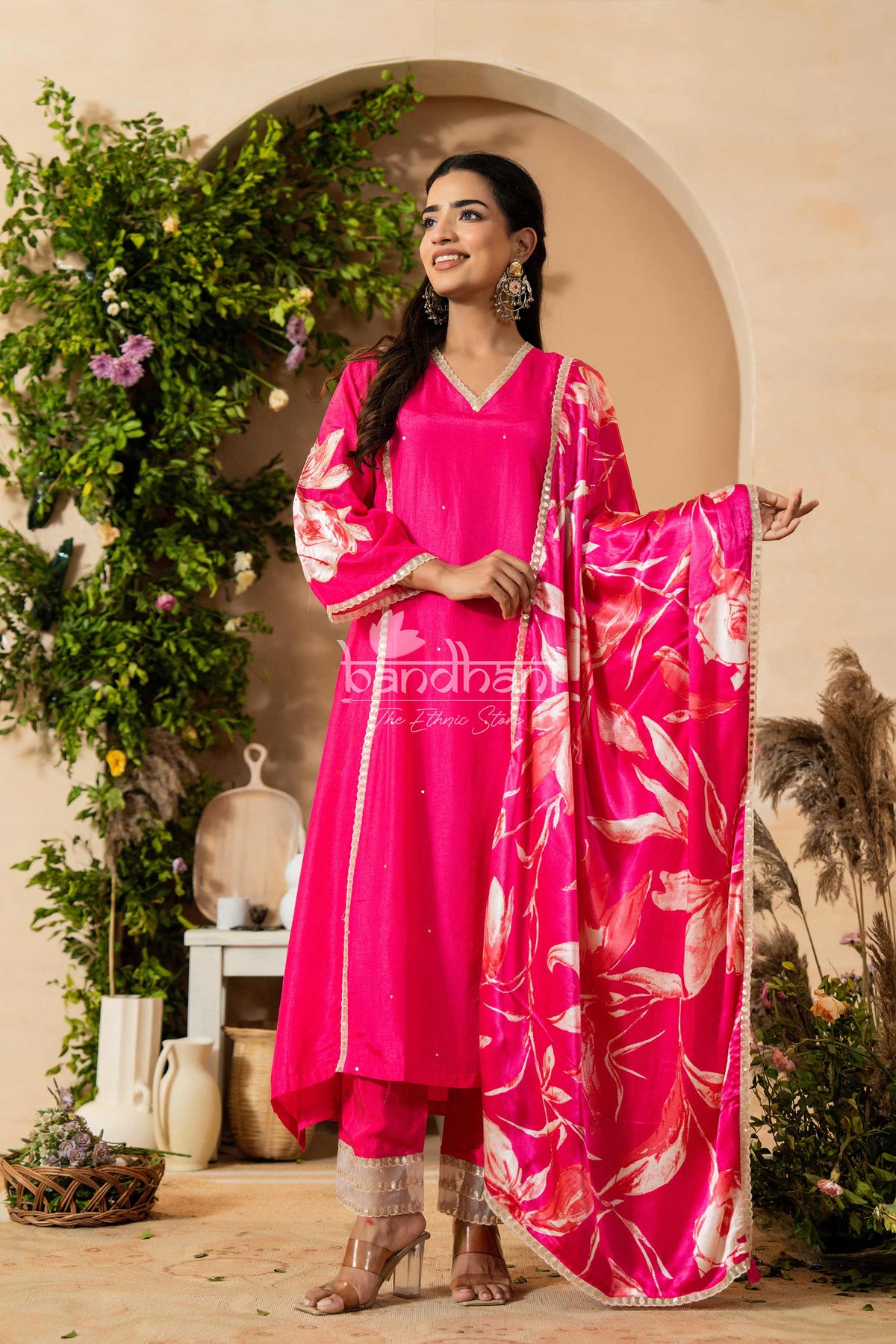 Woman in a bright pink traditional outfit with floral patterns standing in a decorated indoor setting.