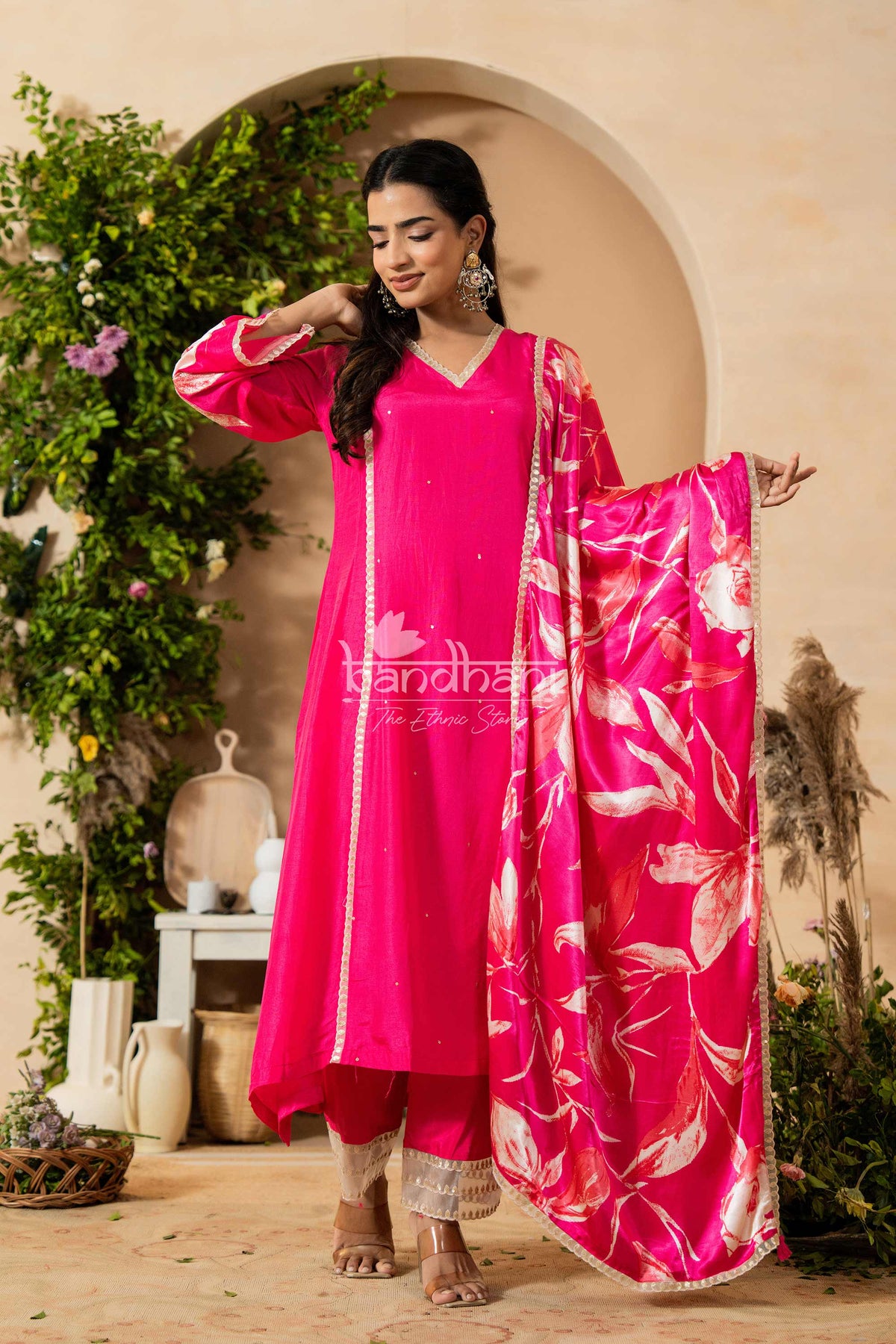 Woman in a bright pink traditional outfit with a floral dupatta, standing in a decorative outdoor setting.