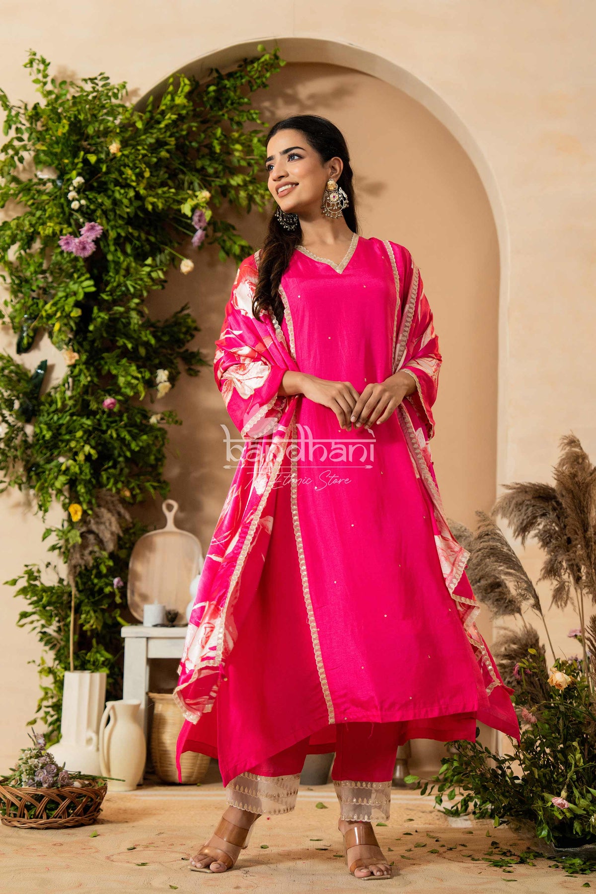 Woman in a bright pink traditional outfit standing in a decorated outdoor setting with 'Bhandari' branding.