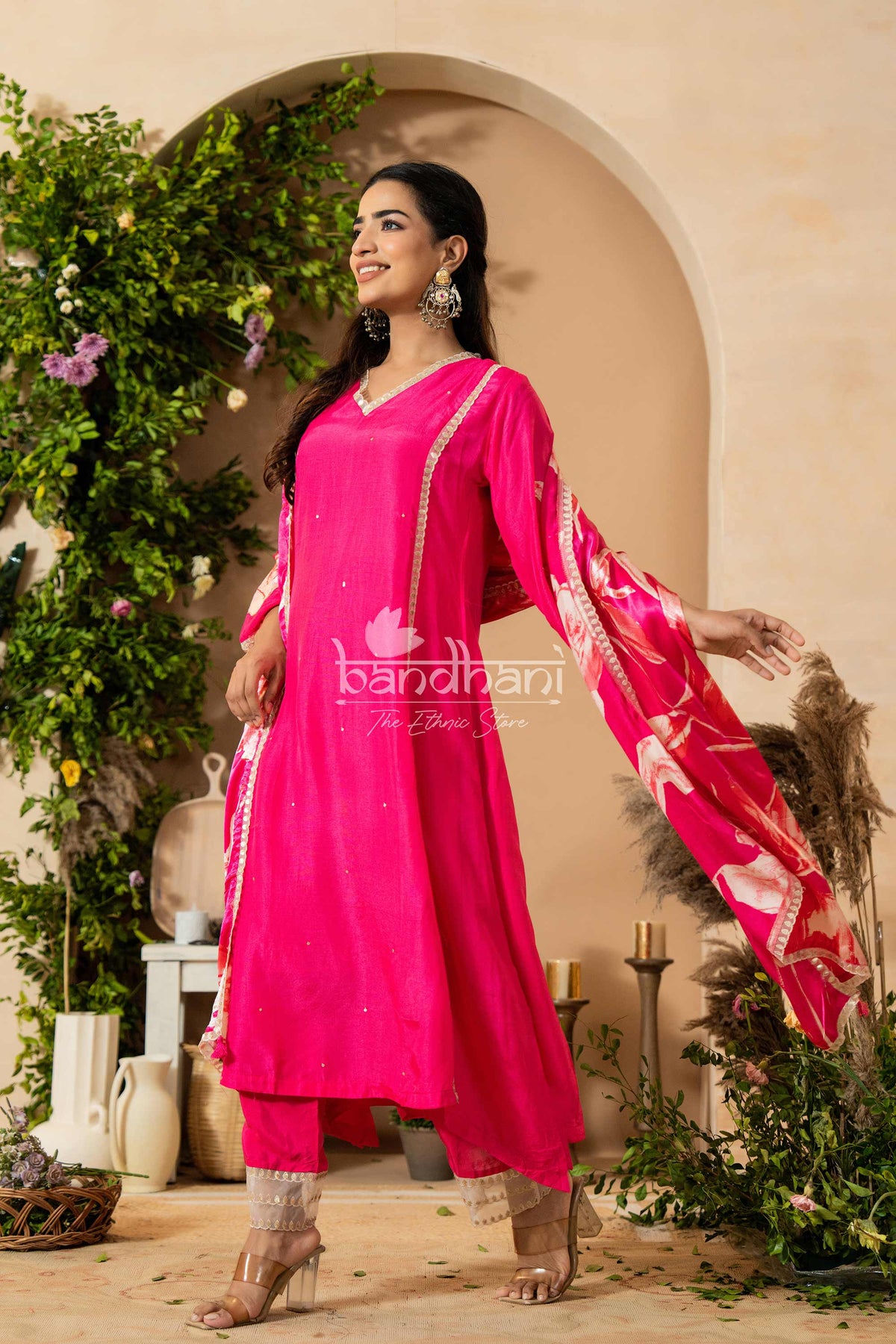 Woman in a bright pink traditional outfit with a floral design, standing in a decorated indoor setting.