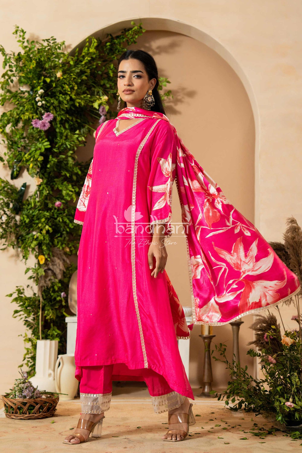 Woman in a bright pink traditional outfit with floral patterns standing in a decorated indoor setting.