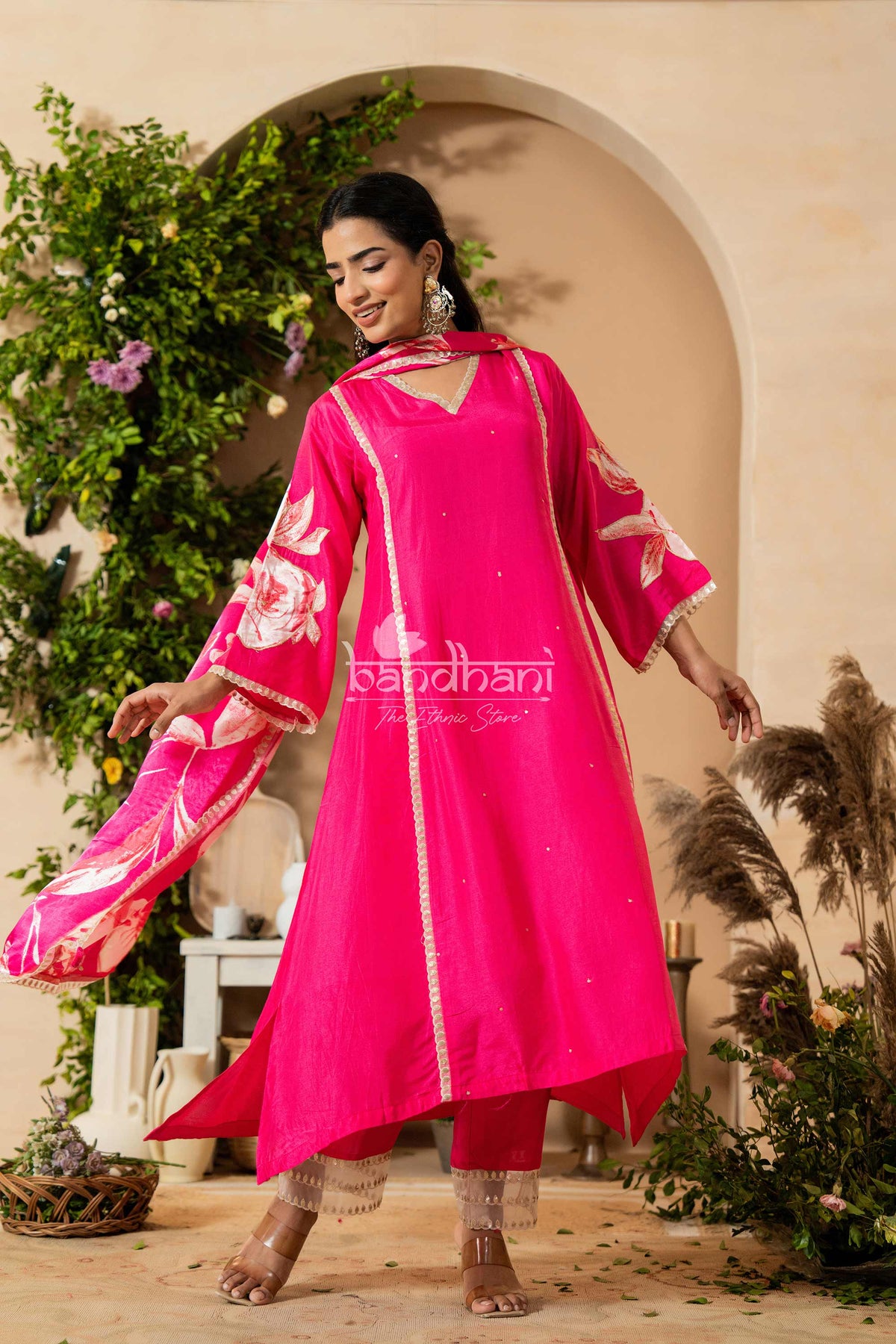 Woman in a bright pink traditional outfit with floral patterns, standing in an outdoor setting with plants and archway.