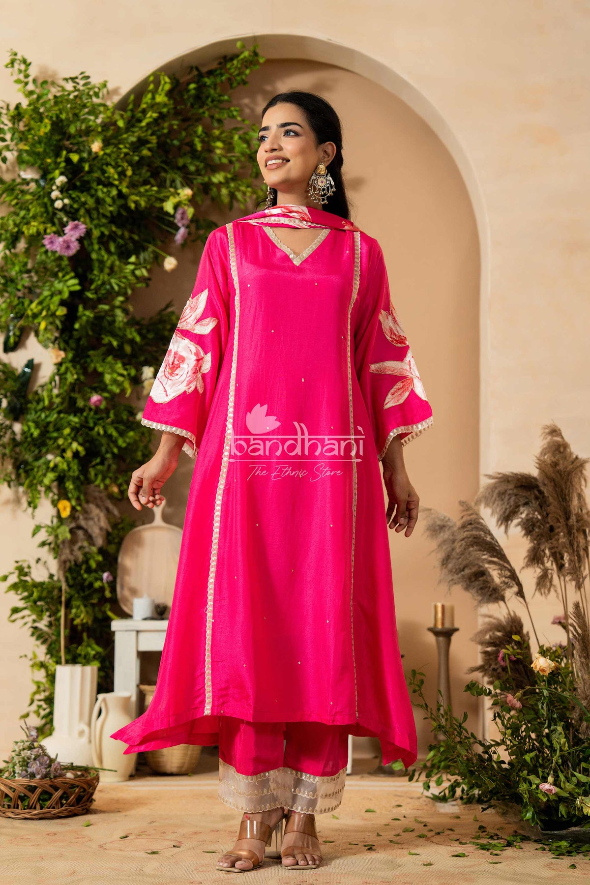 Woman wearing a bright pink traditional outfit with white patterns, standing in a decorative setting.
