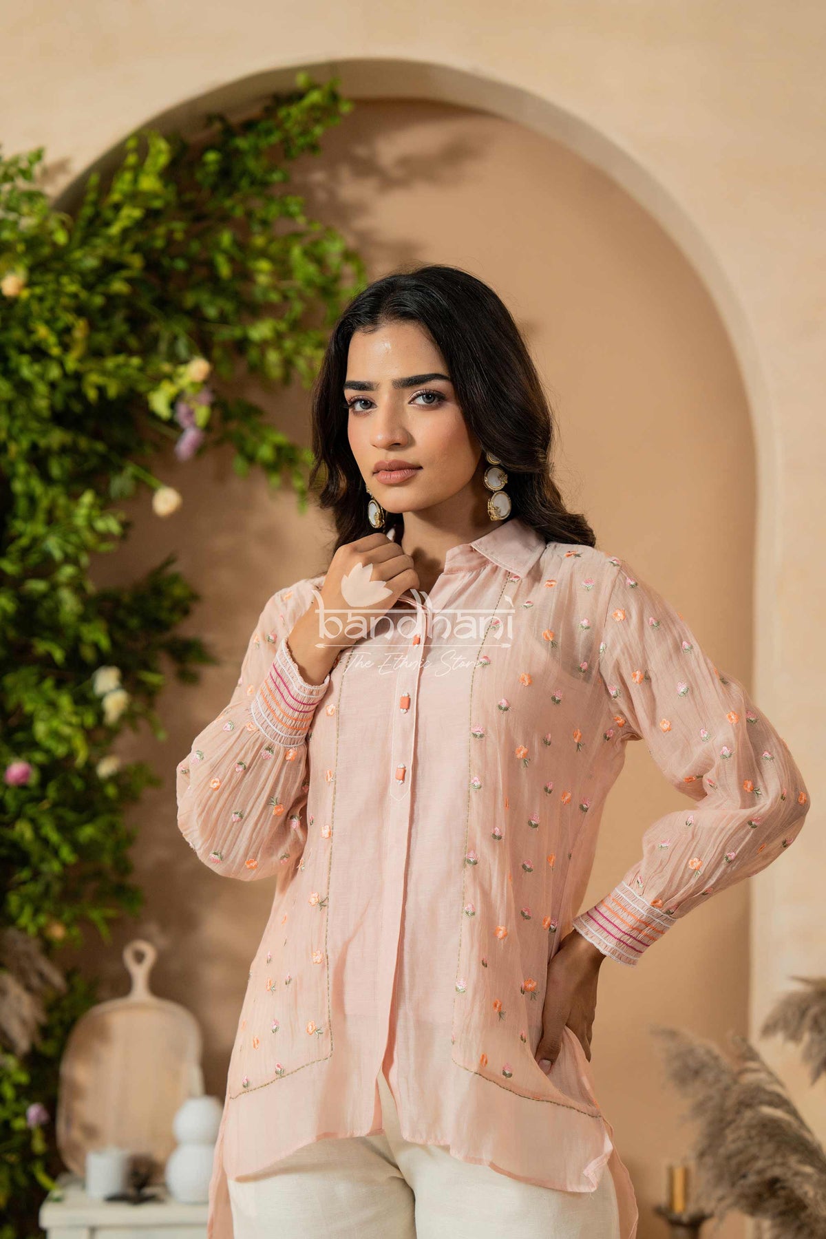 Woman wearing a light pink traditional outfit with intricate designs, standing against a decorative background.