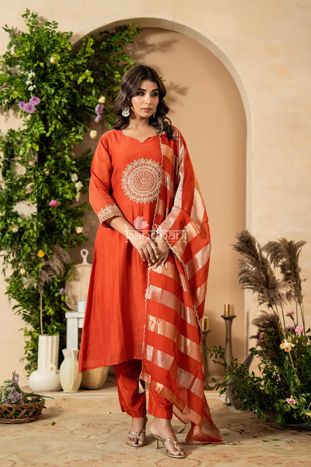 Woman in a red traditional outfit with a decorative pattern, standing in a decorated indoor setting.