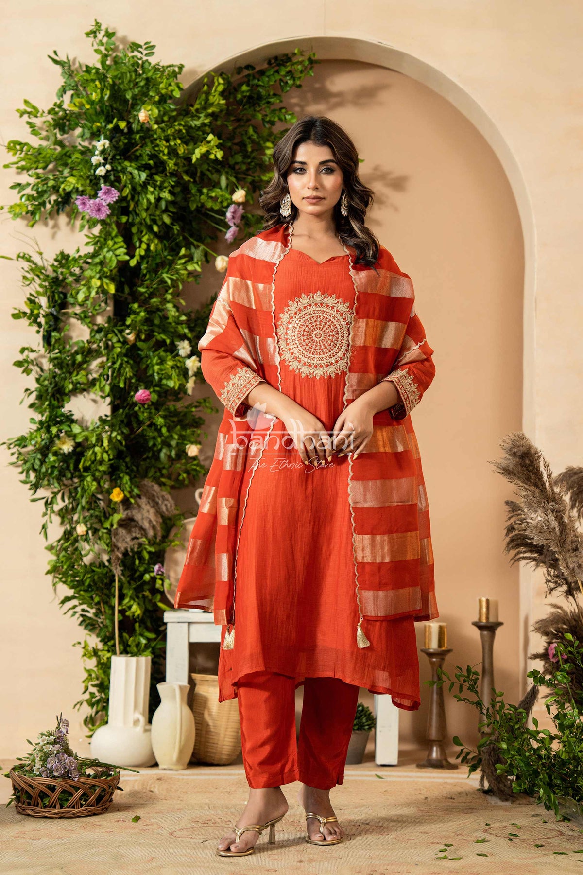 Woman in a red and orange traditional outfit standing in a decorated indoor setting with plants and candles.