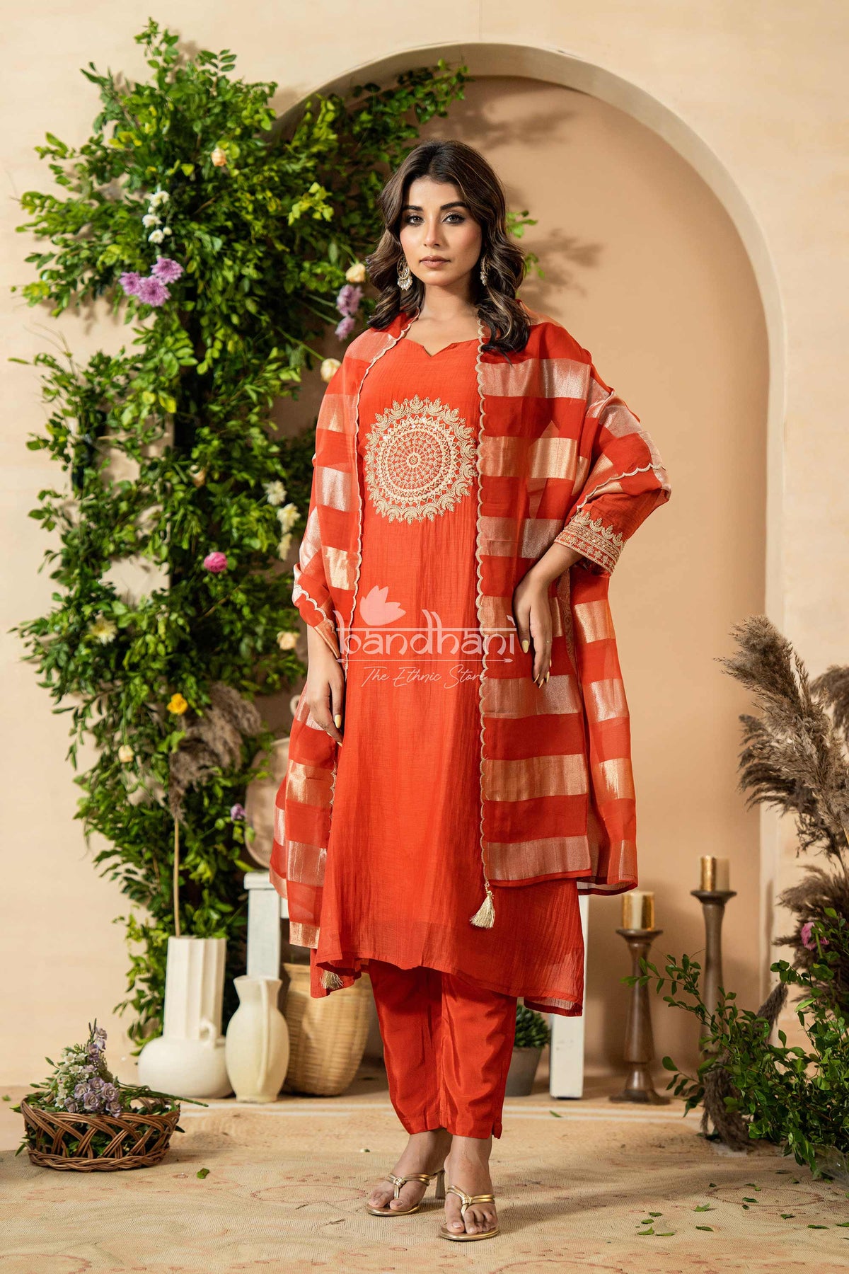 Woman in a red traditional outfit standing in a decorated indoor setting with plants and candles.