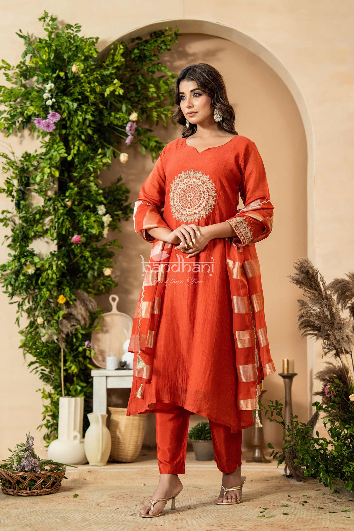 Woman in a red traditional outfit standing in a decorated indoor setting with plants and decor.