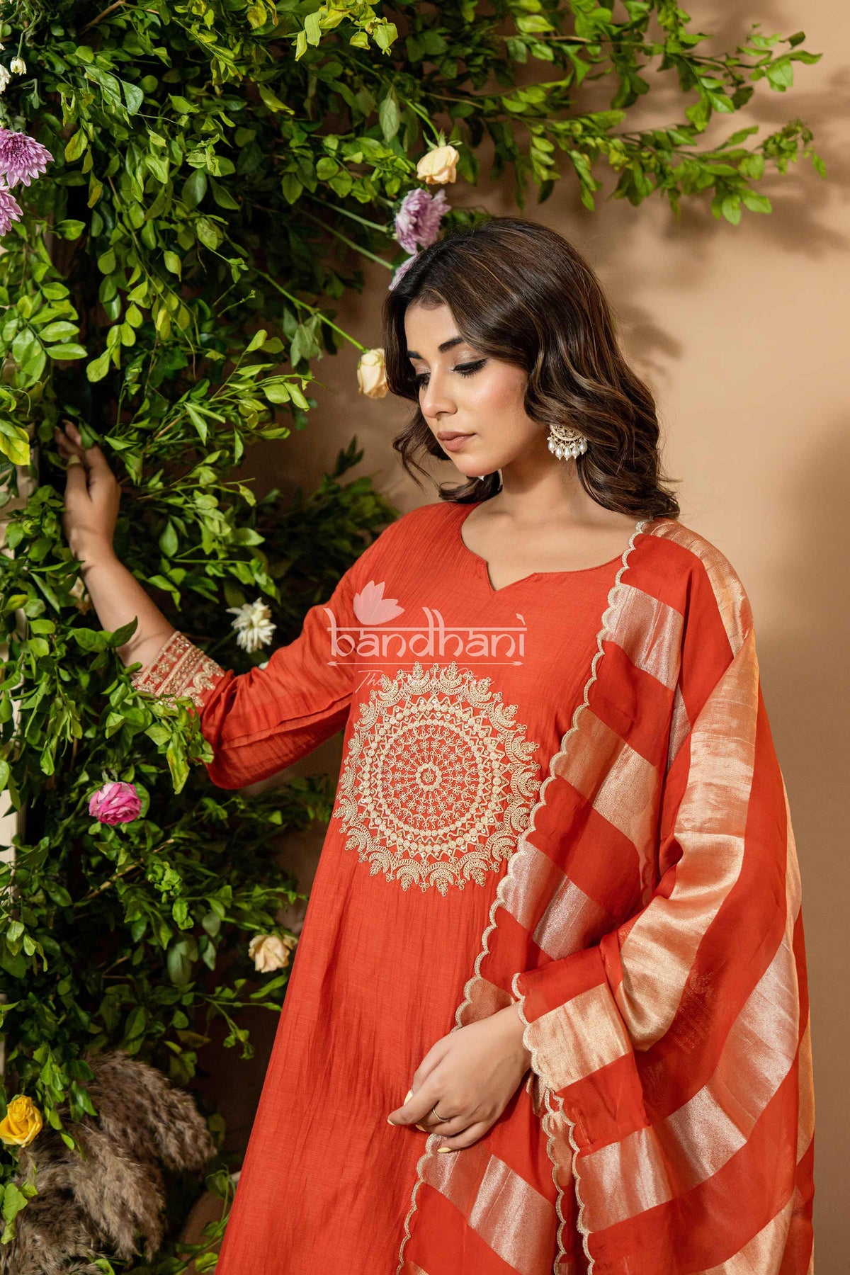 Woman wearing a red traditional outfit with intricate patterns, standing against a green leafy background.