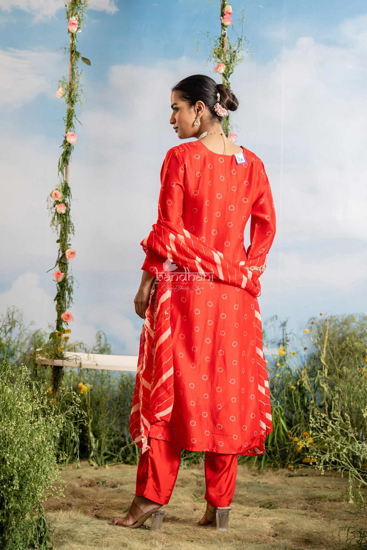 Woman in a red traditional outfit standing outdoors with greenery and a blue sky in the background.
