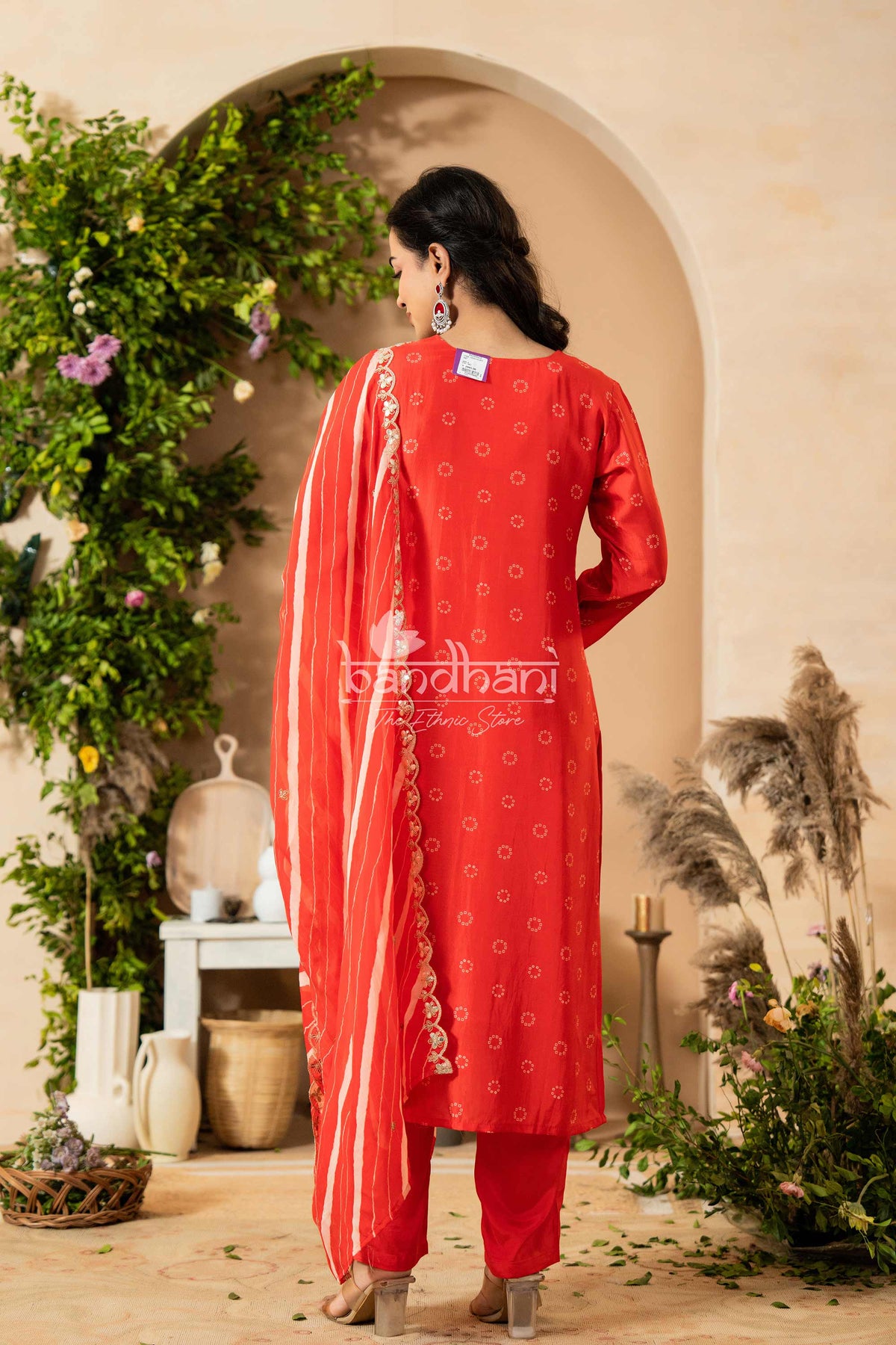 Woman in a red traditional outfit standing in a decorative setting with plants and archway.