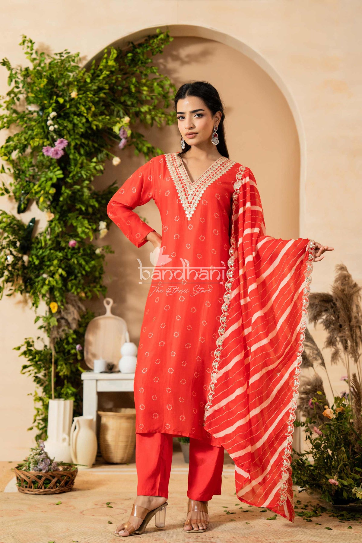 Woman in a red traditional outfit with a dupatta, standing in a decorated indoor setting.
