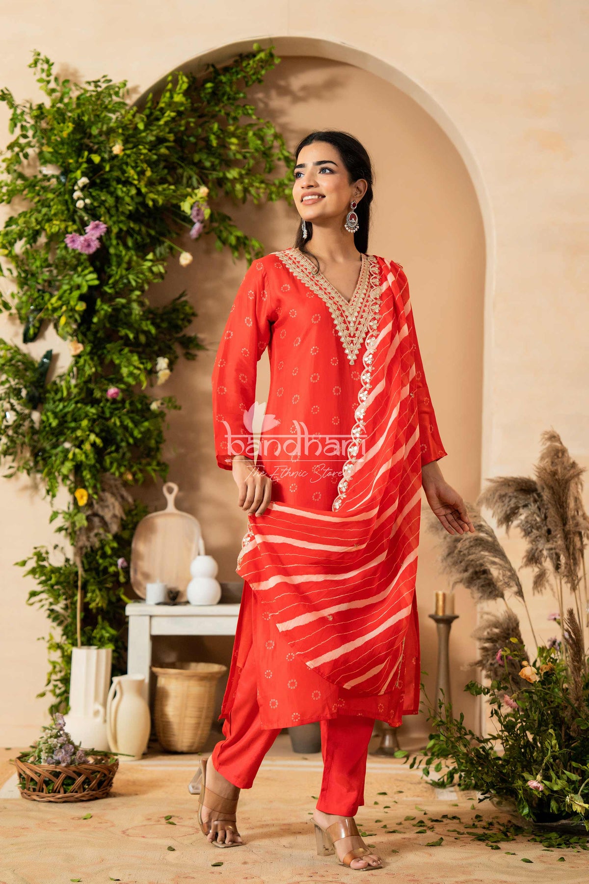 Woman in a red traditional outfit standing in a decorated indoor setting with plants and decor.
