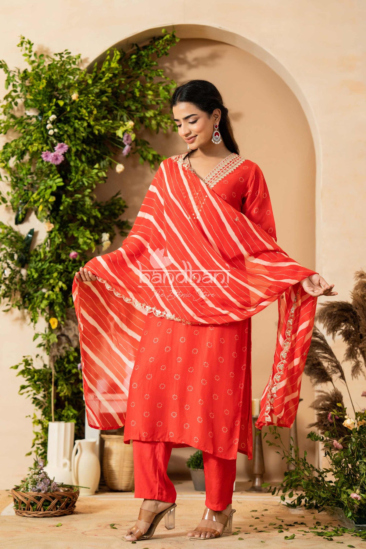 Woman in a red traditional outfit with a matching dupatta, standing in a decorated outdoor setting.