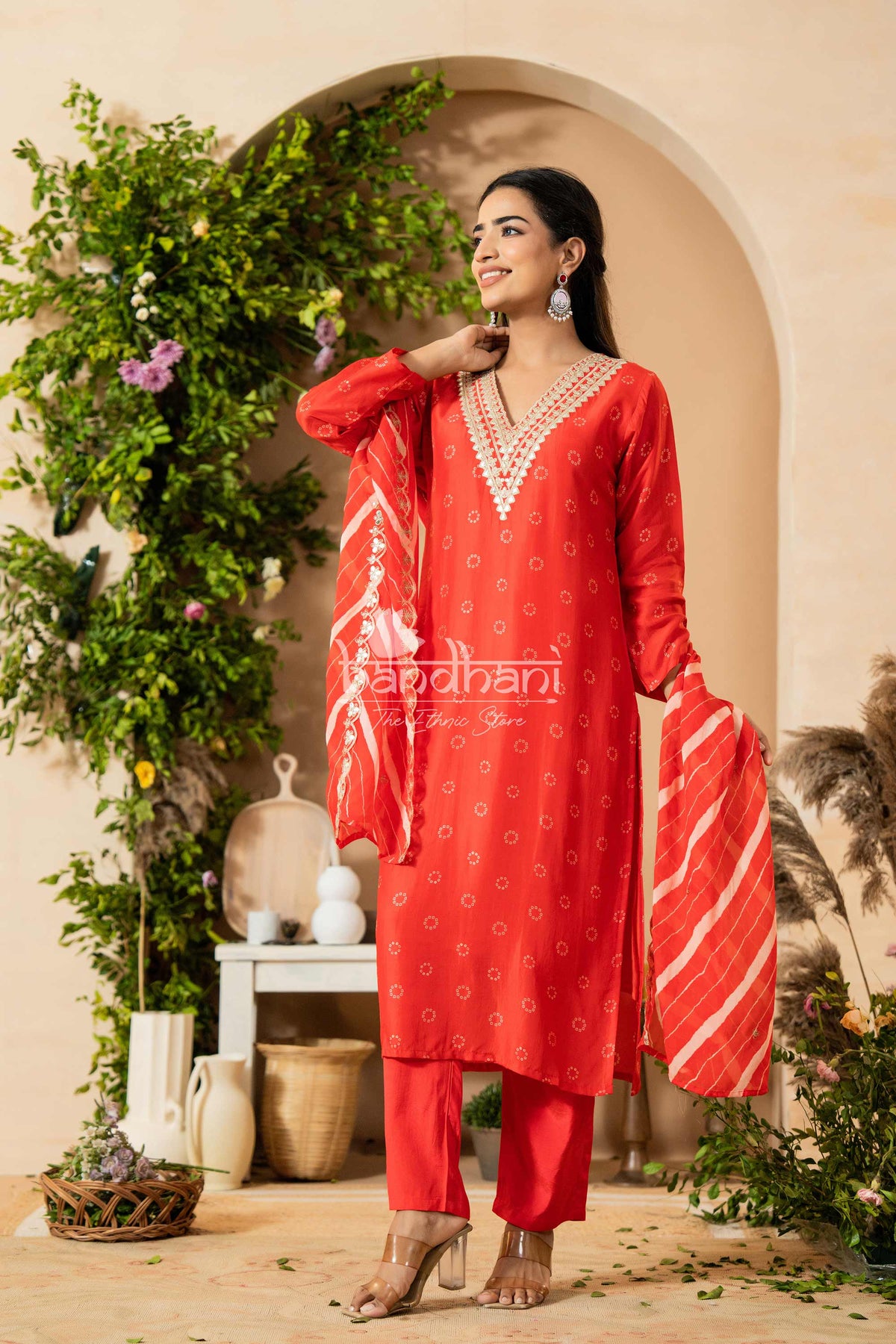 Woman wearing a red traditional outfit with intricate patterns, standing in a decorative setting with plants and a white arch.