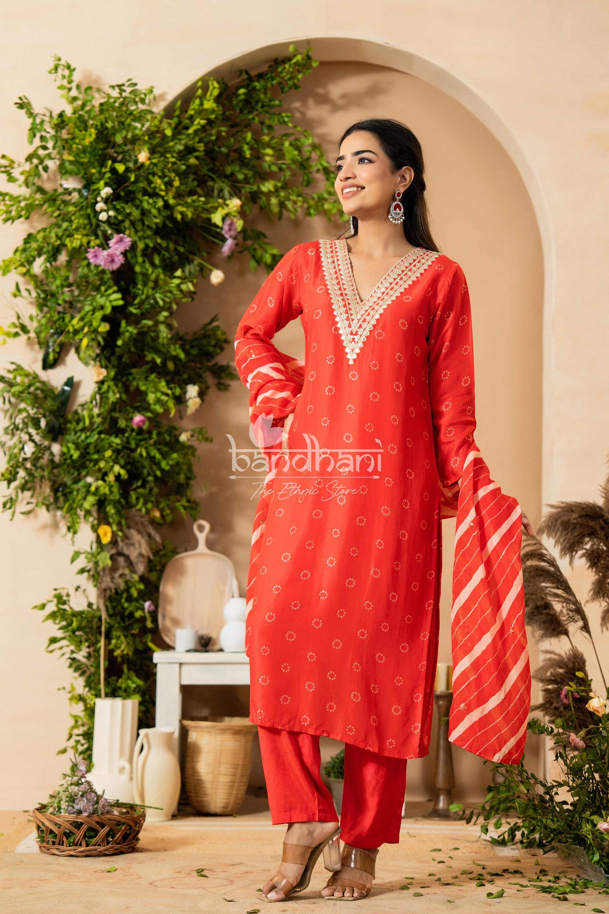 Woman wearing a red traditional outfit with intricate patterns, standing in a decorative indoor setting.