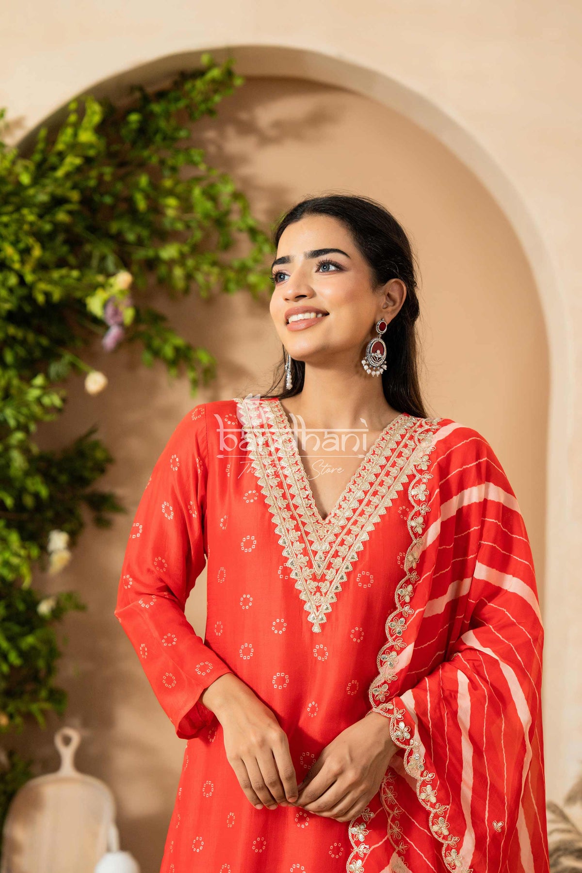 Woman wearing a red traditional outfit with intricate white embroidery, standing against a decorative background.