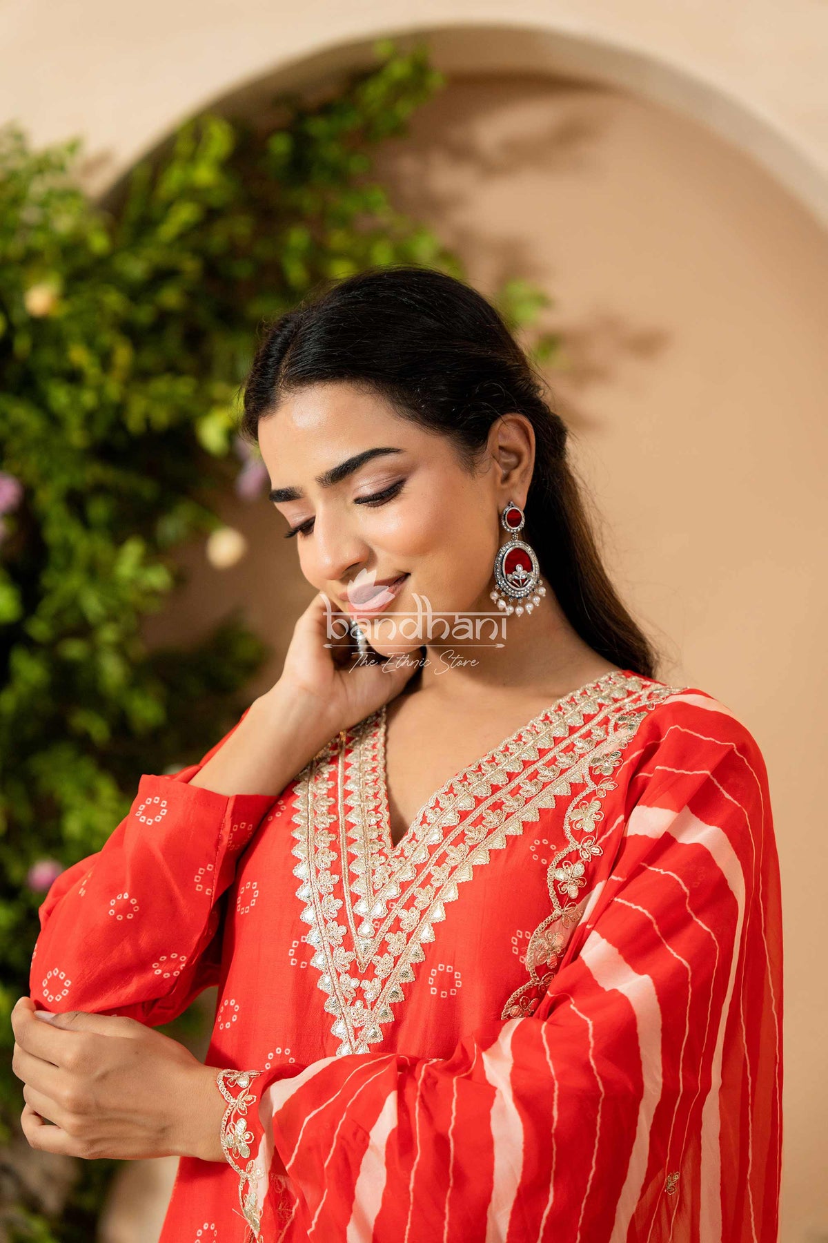 Woman wearing a red traditional outfit with intricate designs, standing against a beige wall with greenery.