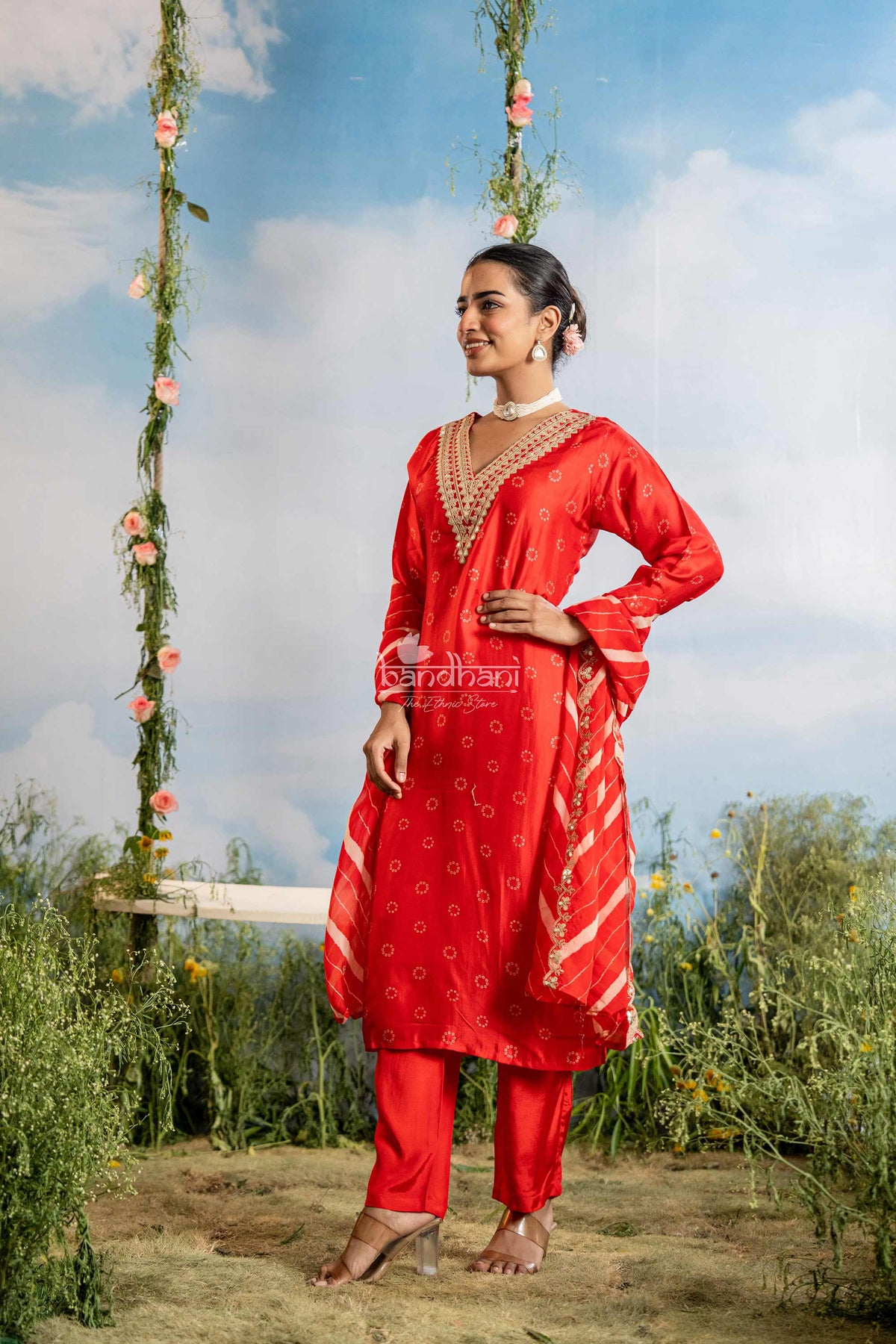 Woman in a red traditional outfit standing outdoors with greenery and a blue sky.