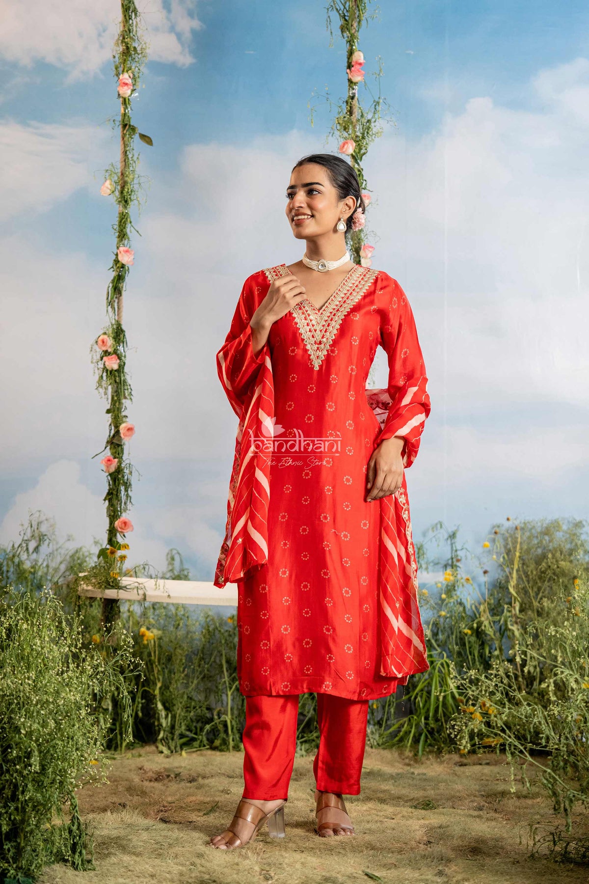 Woman in a red traditional outfit standing on a grassy area with a swing and floral decorations in the background.