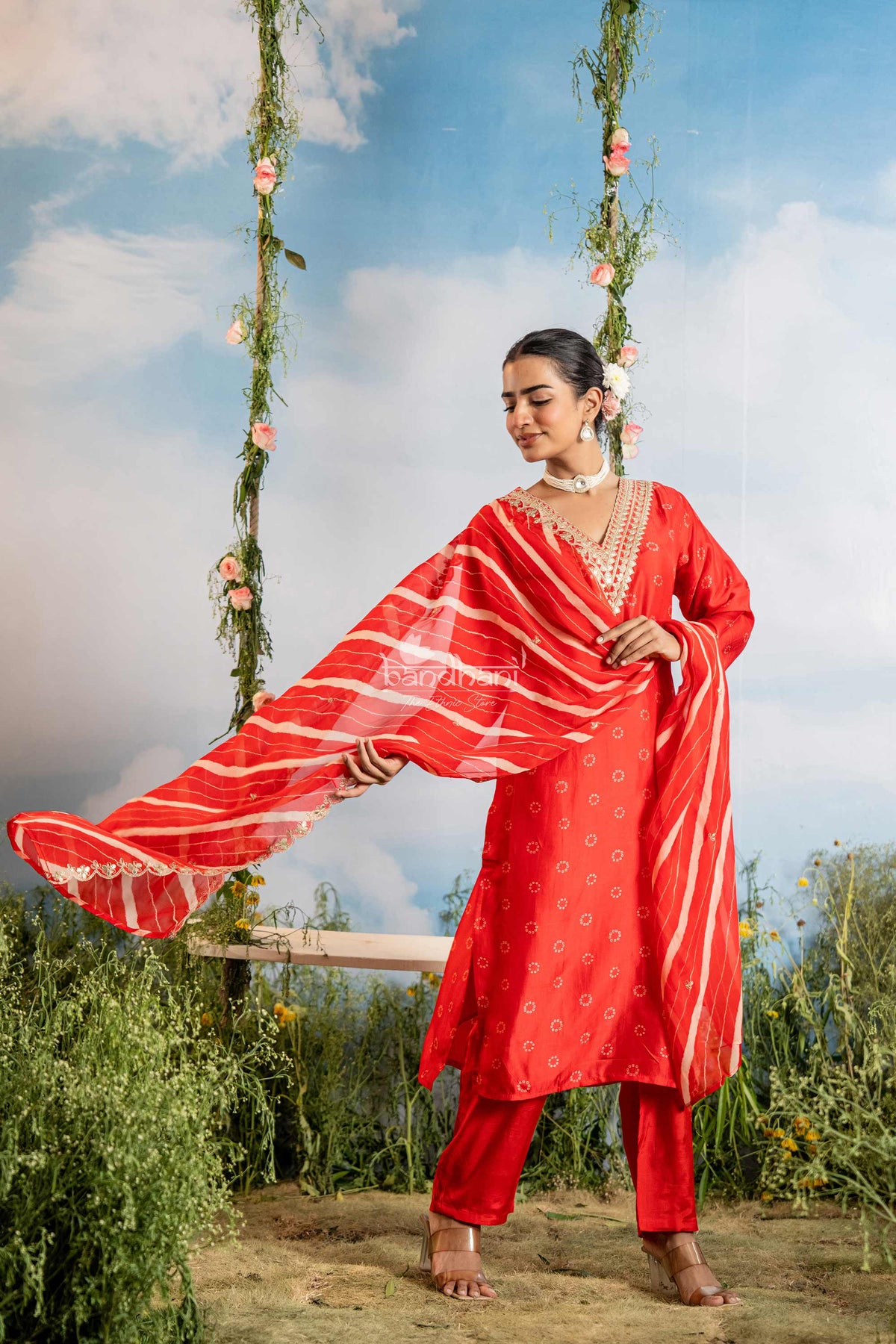 Woman in a red traditional outfit sitting on a swing with floral decorations against a blue sky.