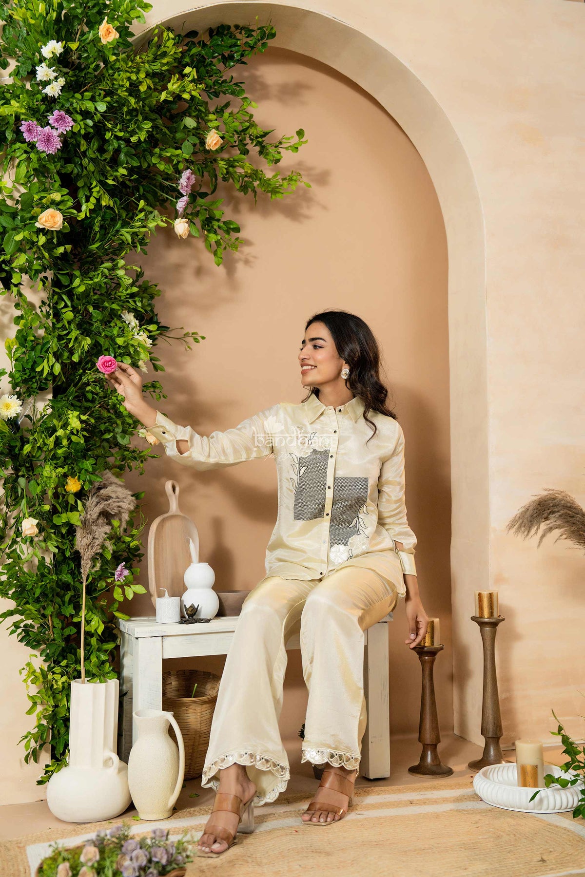 Woman in a cream traditional outfit sitting in a decorated room with flowers and decor.
