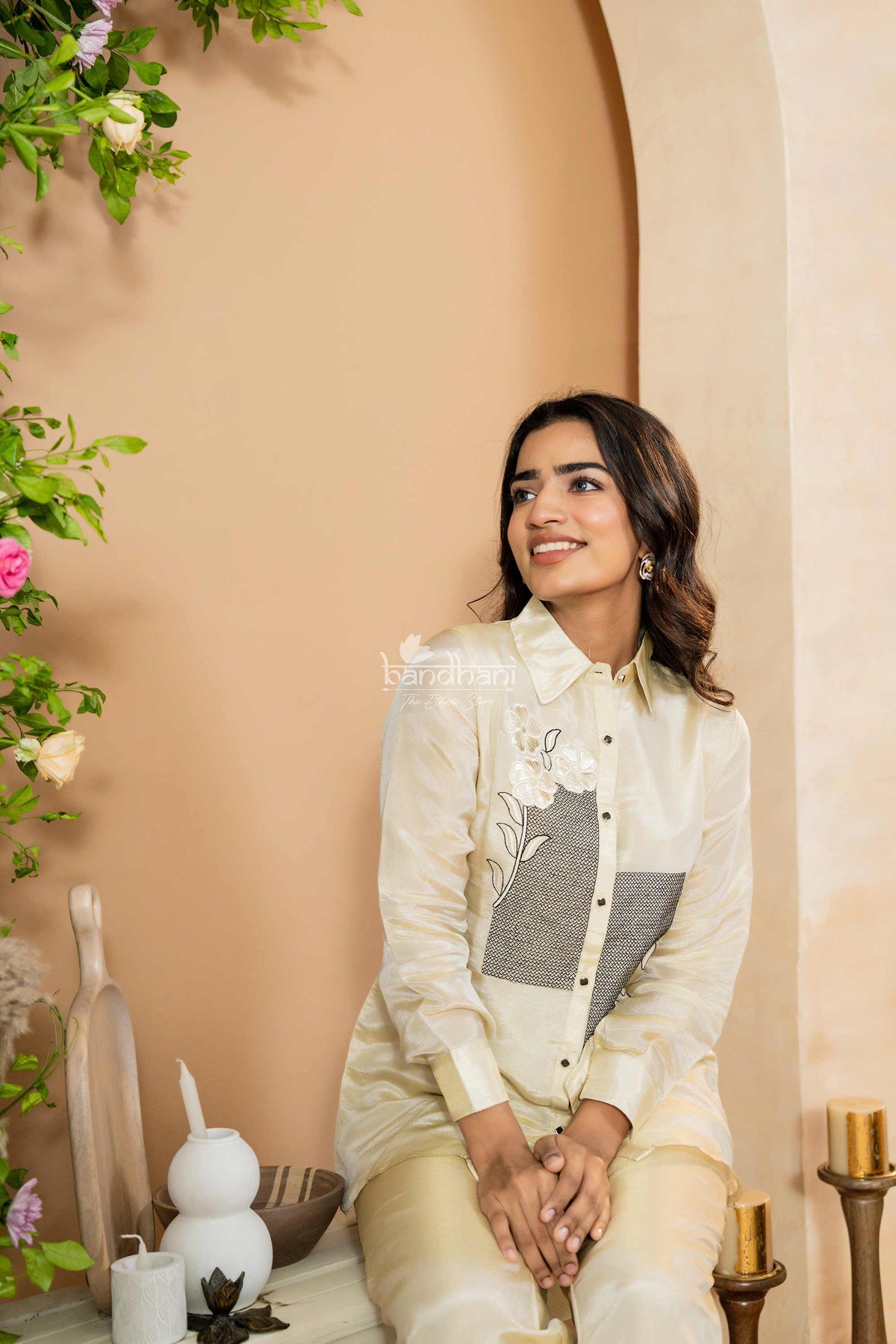Woman in a white traditional outfit sitting in a decorated indoor setting with plants and candles.