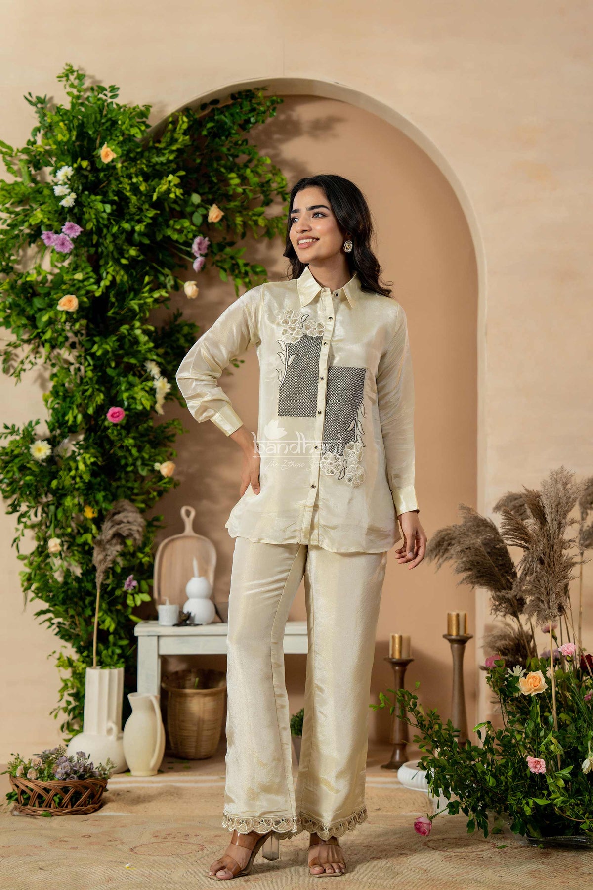 Woman in a traditional outfit standing in a decorated room with plants and flowers.