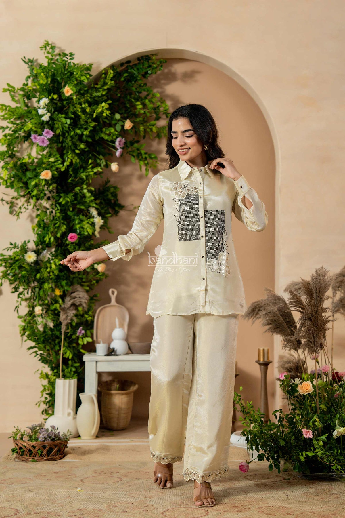 Woman in traditional outfit standing in a decorated indoor setting with flowers and plants.