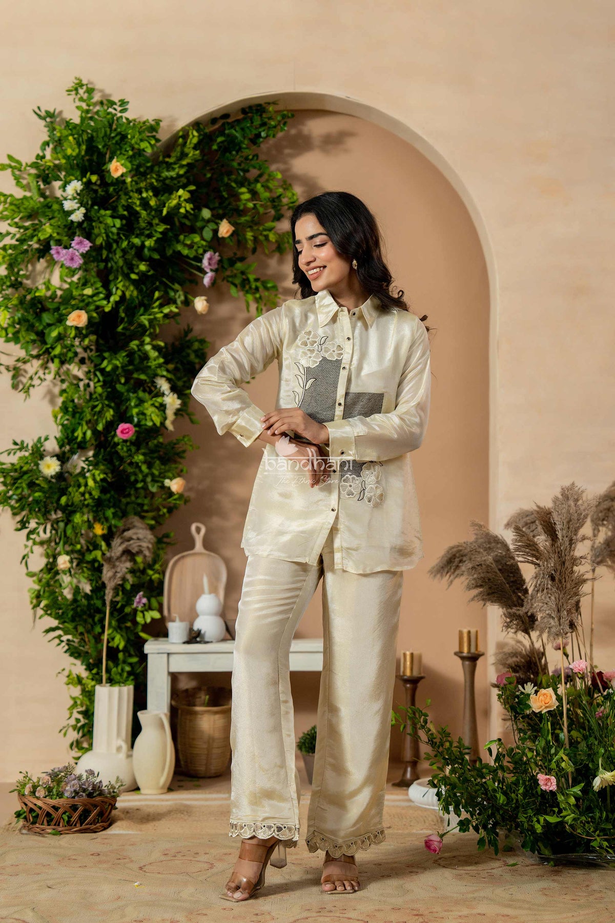 Woman in a cream embroidered outfit standing in a decorated indoor setting with plants and flowers.