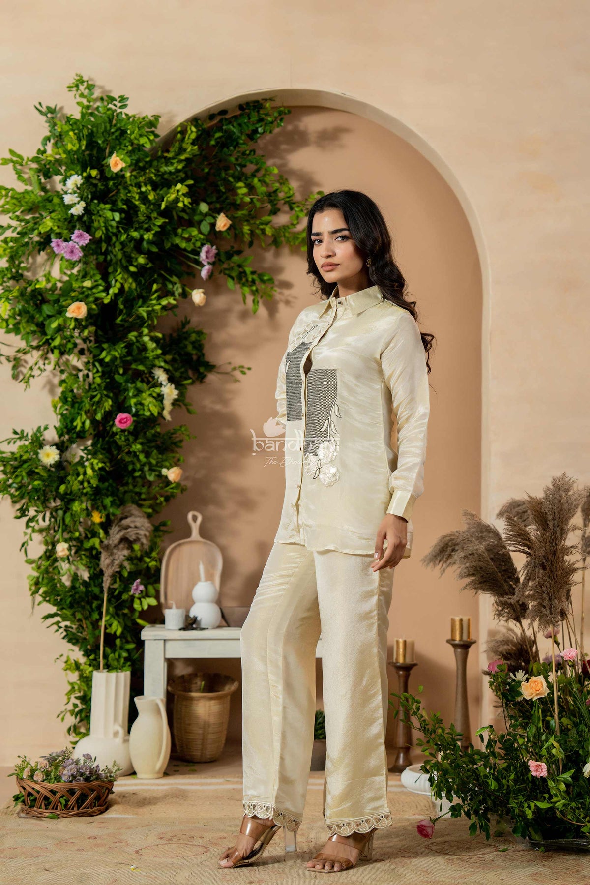 Woman in a light-colored outfit standing in a decorated indoor setting with plants and flowers.