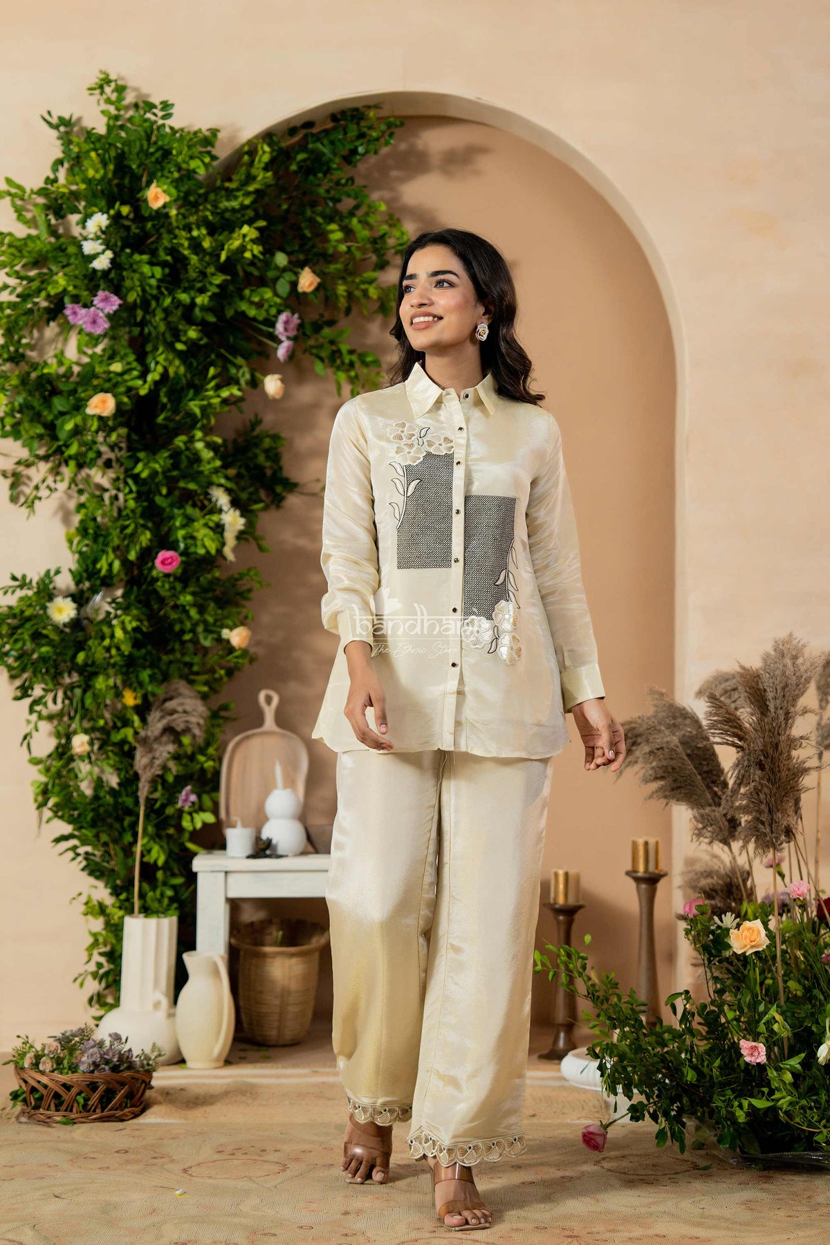 Woman in a stylish outfit standing in a decorated outdoor setting with plants and flowers.