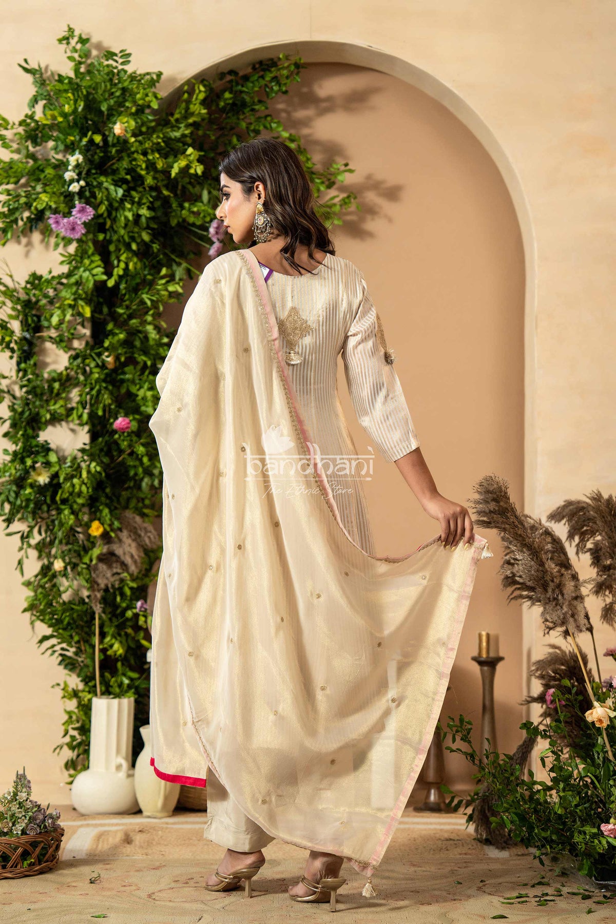 Woman in a white traditional outfit standing in a decorated indoor setting with plants and flowers.