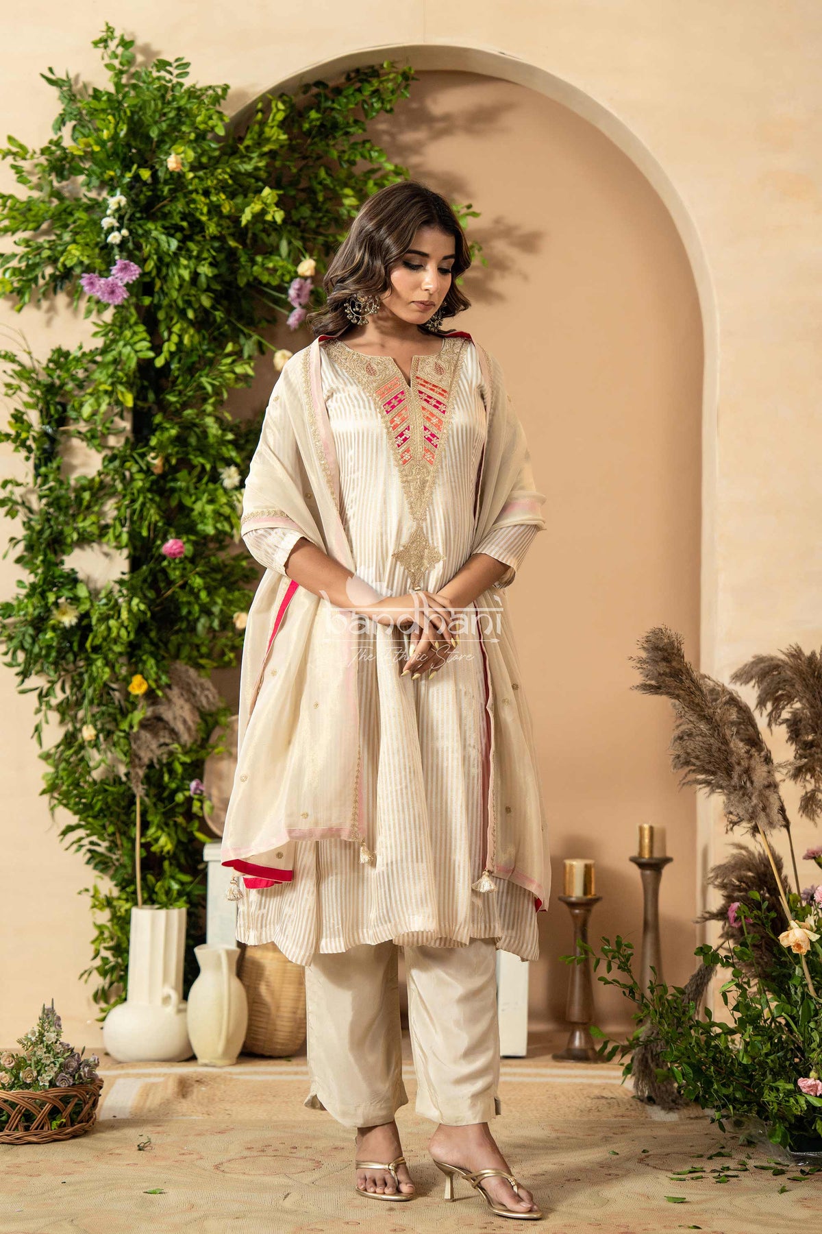 Woman in a traditional outfit standing in a decorated indoor setting with plants and candles.